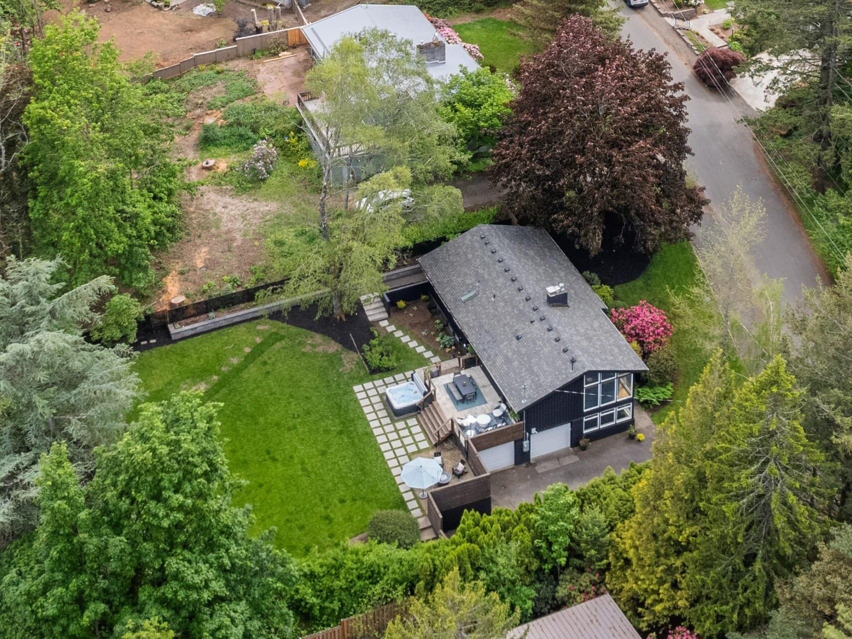 Aerial view of a house with a dark roof surrounded by lush green trees. The property features a large grassy yard, a patio with outdoor furniture, and a neighboring road. The landscape includes both manicured and natural vegetation.