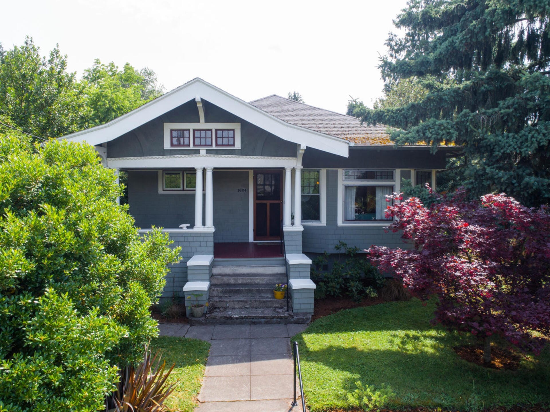 A cozy gray bungalow with a covered porch and white trim, surrounded by lush greenery and mature trees. A concrete path leads to the entrance, and colorful shrubs add charm to the front yard.