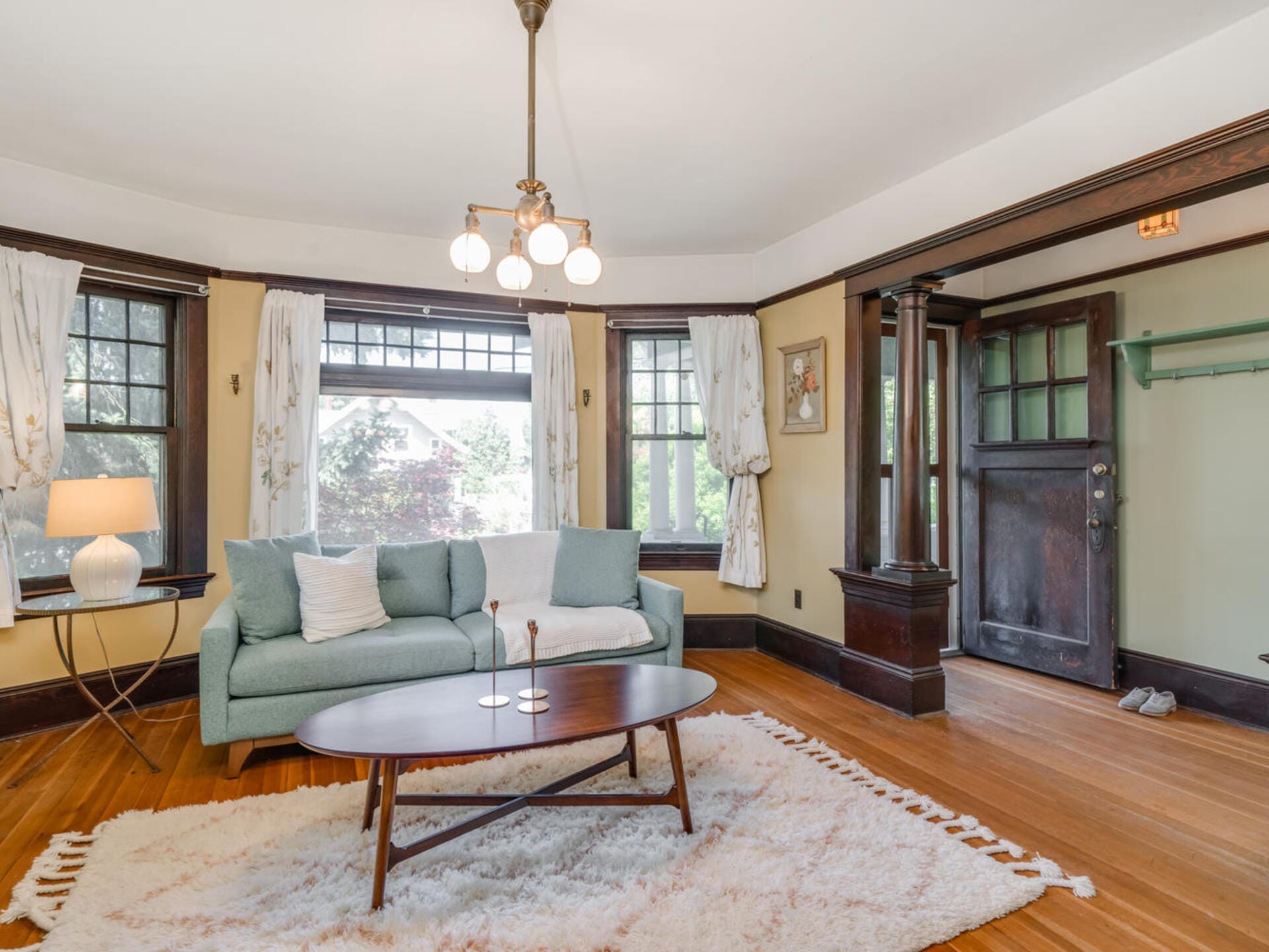 A cozy living room featuring a mint green sofa with white pillows, a wooden coffee table on a fluffy white rug, and large windows with light curtains. The space is adorned with a ceiling light fixture and a dark wooden entry door.