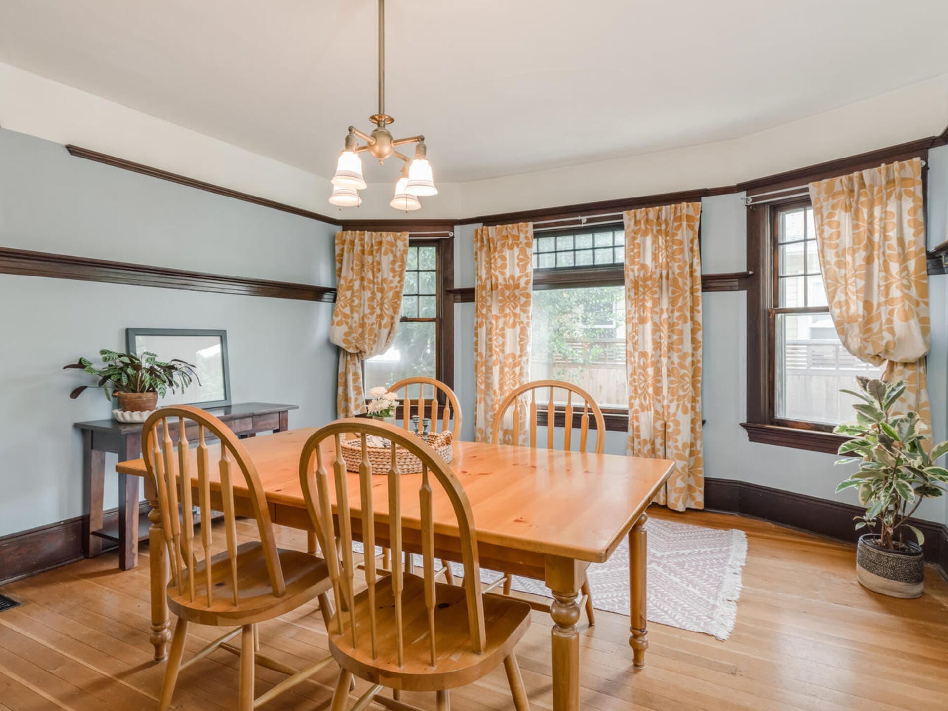 A bright dining room with a wooden table and four chairs. The room features light blue walls, floral-patterned curtains, plants, and a woven rug. A hanging light fixture illuminates the space, which has large windows letting in natural light.