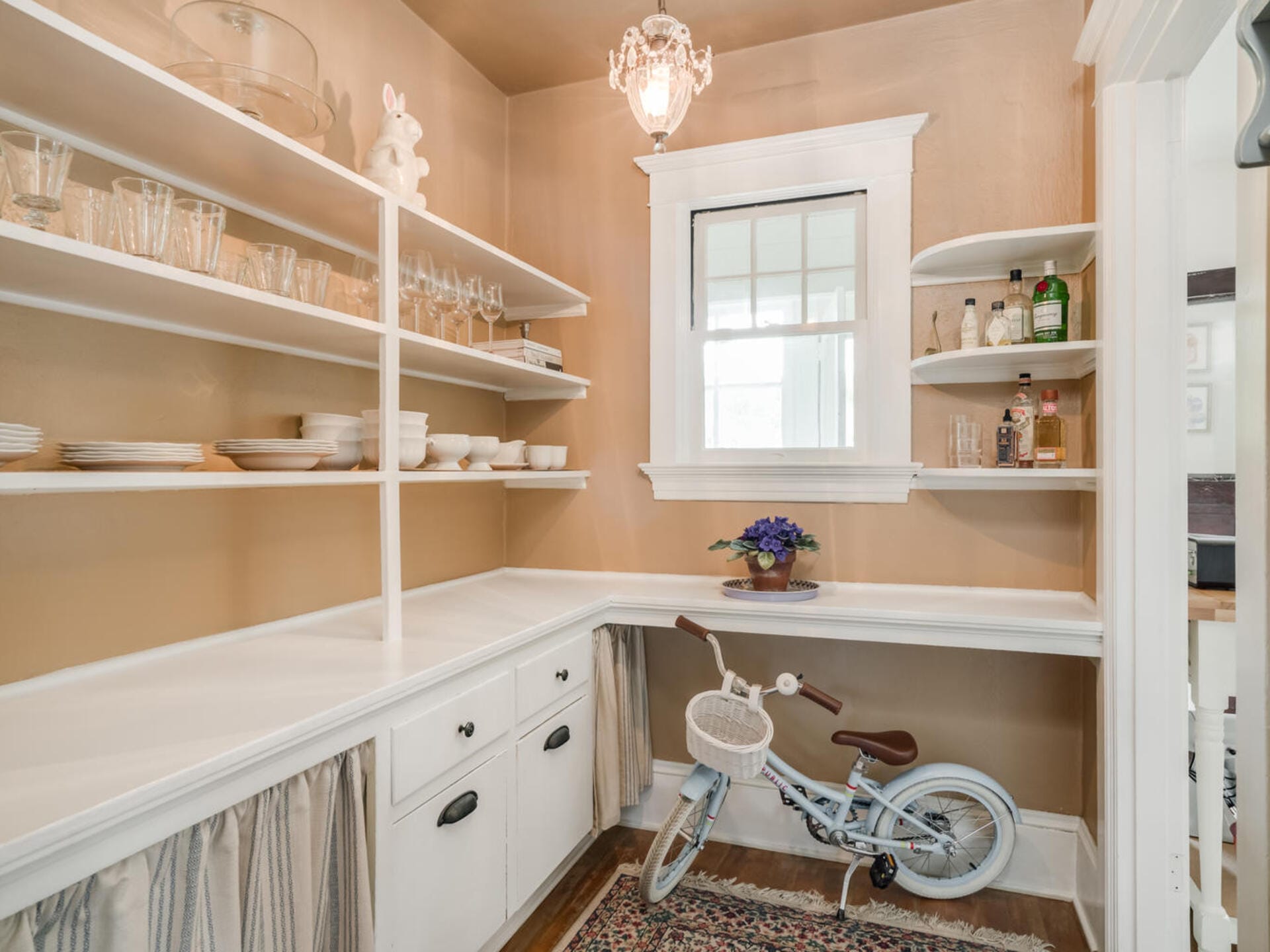 A neatly organized pantry with white shelves displaying glassware and dishes. A vintage-style chandelier hangs overhead. A small window lets in natural light. On the floor, a blue bicycle rests beneath the counter. A floral rug adds color to the floor.