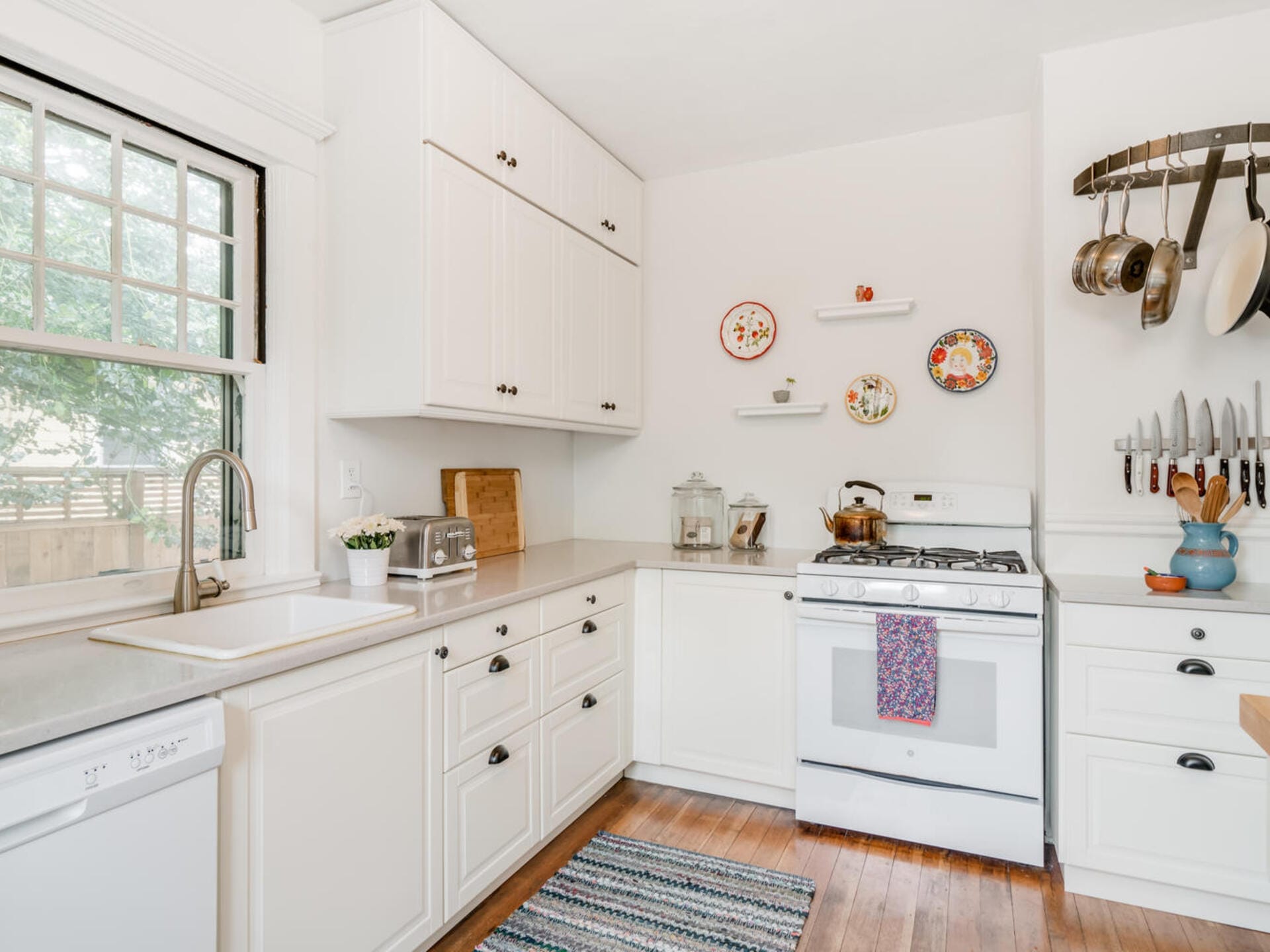 A bright kitchen with white cabinets and a large window. It features a stove with a colorful towel, a knife rack, and hanging pots. Decorative plates adorn the wall above open shelves. A striped rug and toaster are visible on the wooden floor.