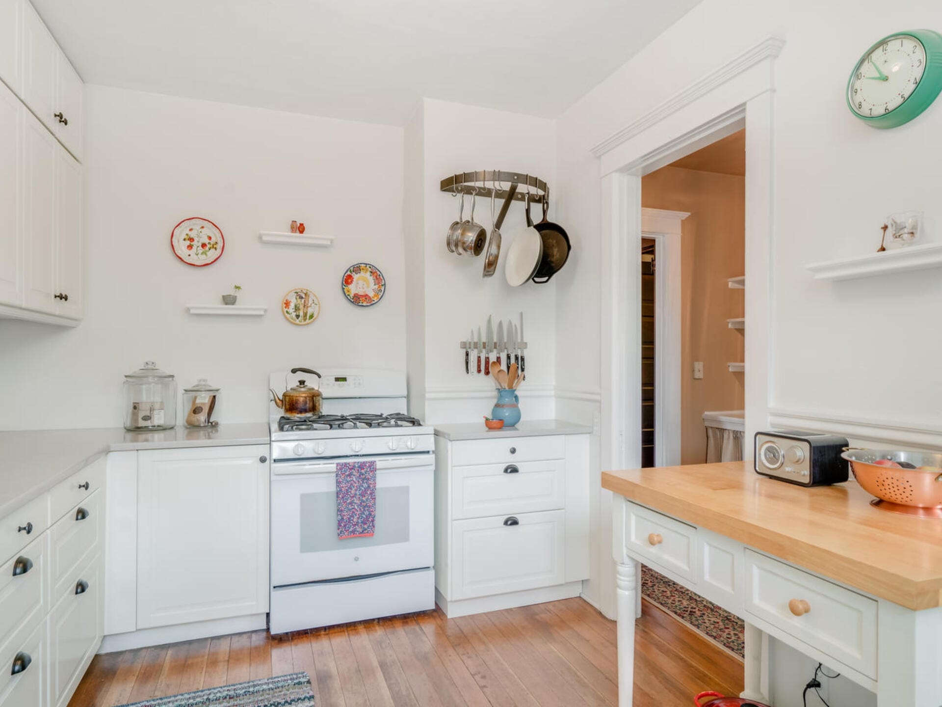 A cozy white kitchen with wooden flooring, featuring a gas stove, white cabinets, and decorative plates on the wall. A wooden island holds a radio and fruit bowl. A utensil rack with pots and pans hangs above the countertop.