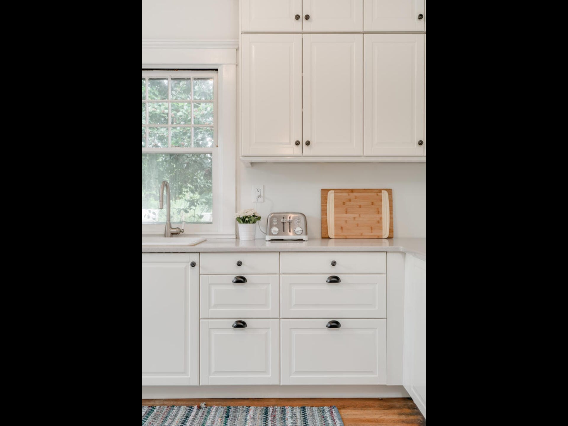 A bright kitchen corner with white cabinets and drawers. A window with a view of greenery, a countertop with a cutting board, small toaster, and a potted plant are visible. A striped rug covers part of the wooden floor.