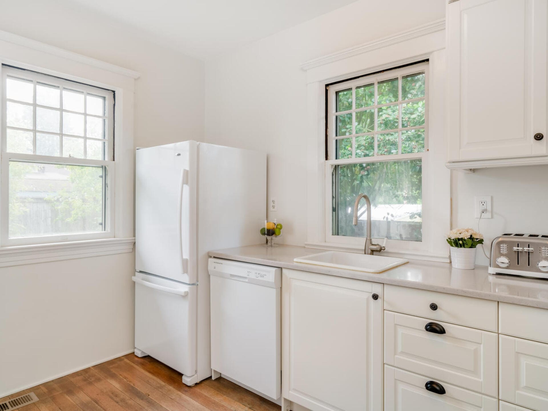 A bright kitchen with white cabinets, a refrigerator, and a dishwasher. Two large windows provide natural light. A potted plant and a bowl of lemons sit on the counter near a stainless steel sink and a toaster.