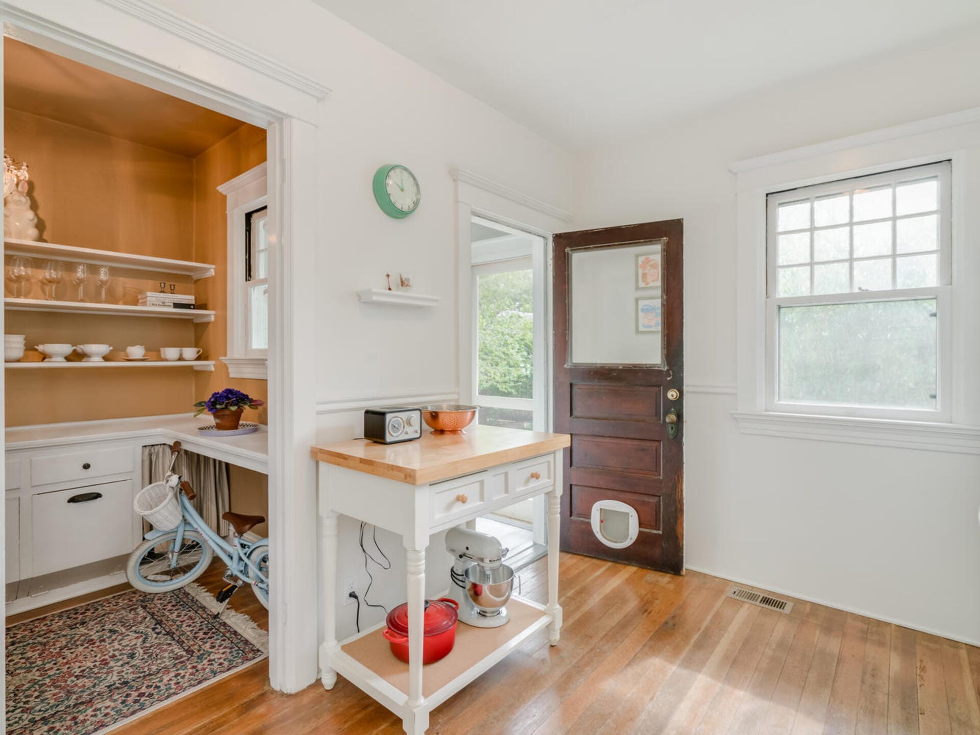 A cozy kitchen features a small wooden island with a mixer, a vintage radio, and a red bowl. An adjacent pantry displays shelves with dishes and a bicycle. Hardwood floors and a cat door in the wooden back door complete the scene.