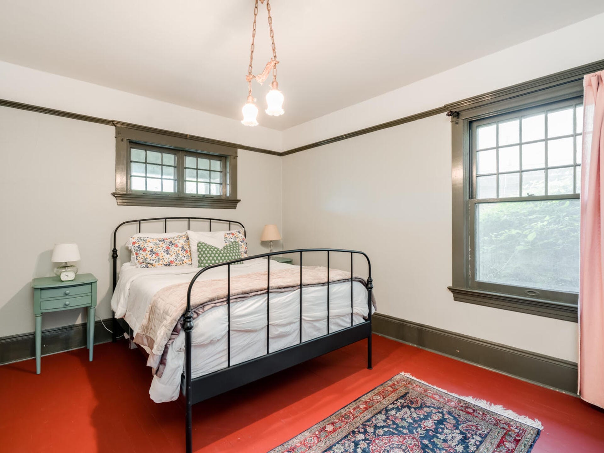 A cozy bedroom featuring a black metal bed with floral and polka dot pillows and a white comforter. There are two windows with dark trim, a small green nightstand with a lamp, and a colorful rug on the red floor. A hanging light fixture illuminates the room.