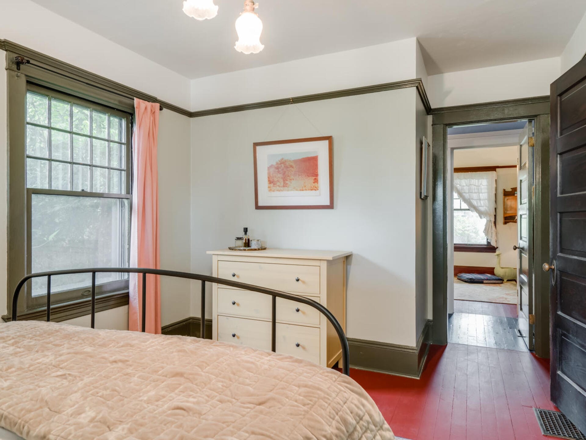A cozy bedroom with a metal-framed bed, beige comforter, and a white dresser. A window with pink curtains is on the left, and a framed artwork hangs above the dresser. A doorway leads to another room with wooden floors. Hanging lights are visible.
