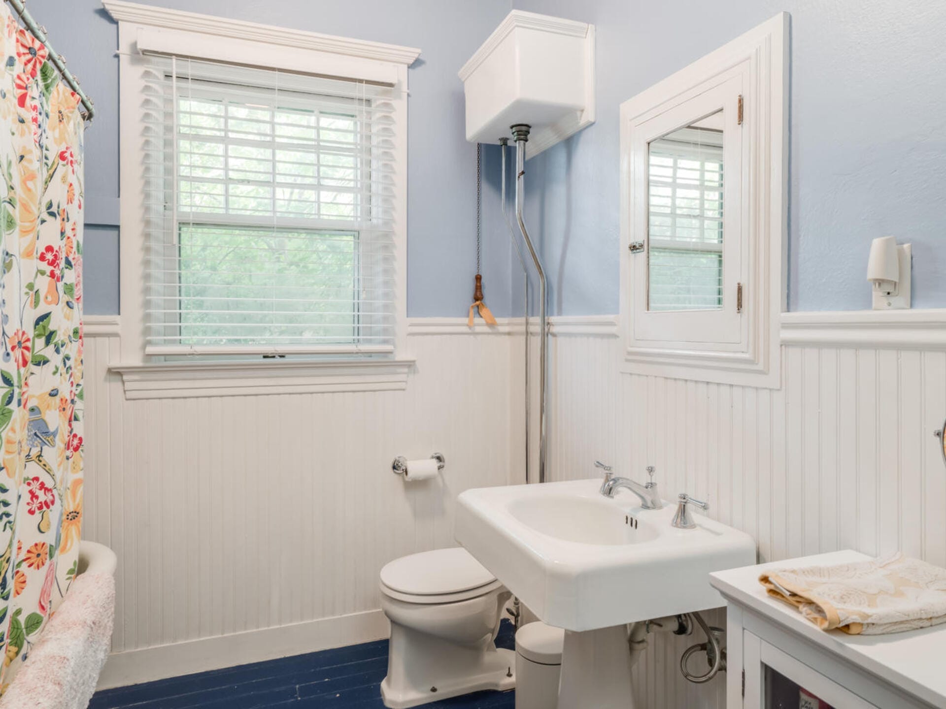 A small bathroom with blue and white walls, featuring a pedestal sink, a mirror, and a toilet with a high tank. A colorful floral shower curtain hangs beside the window, which has white blinds. A white towel sits on a counter.