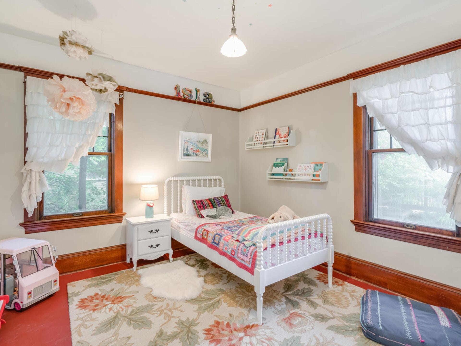 A cozy childrens bedroom featuring a white bed with colorful bedding, a nightstand with a lamp, shelves with books and toys, and two large windows with ruffled curtains. A rug covers the hardwood floor, and a toy vehicle is in the corner.