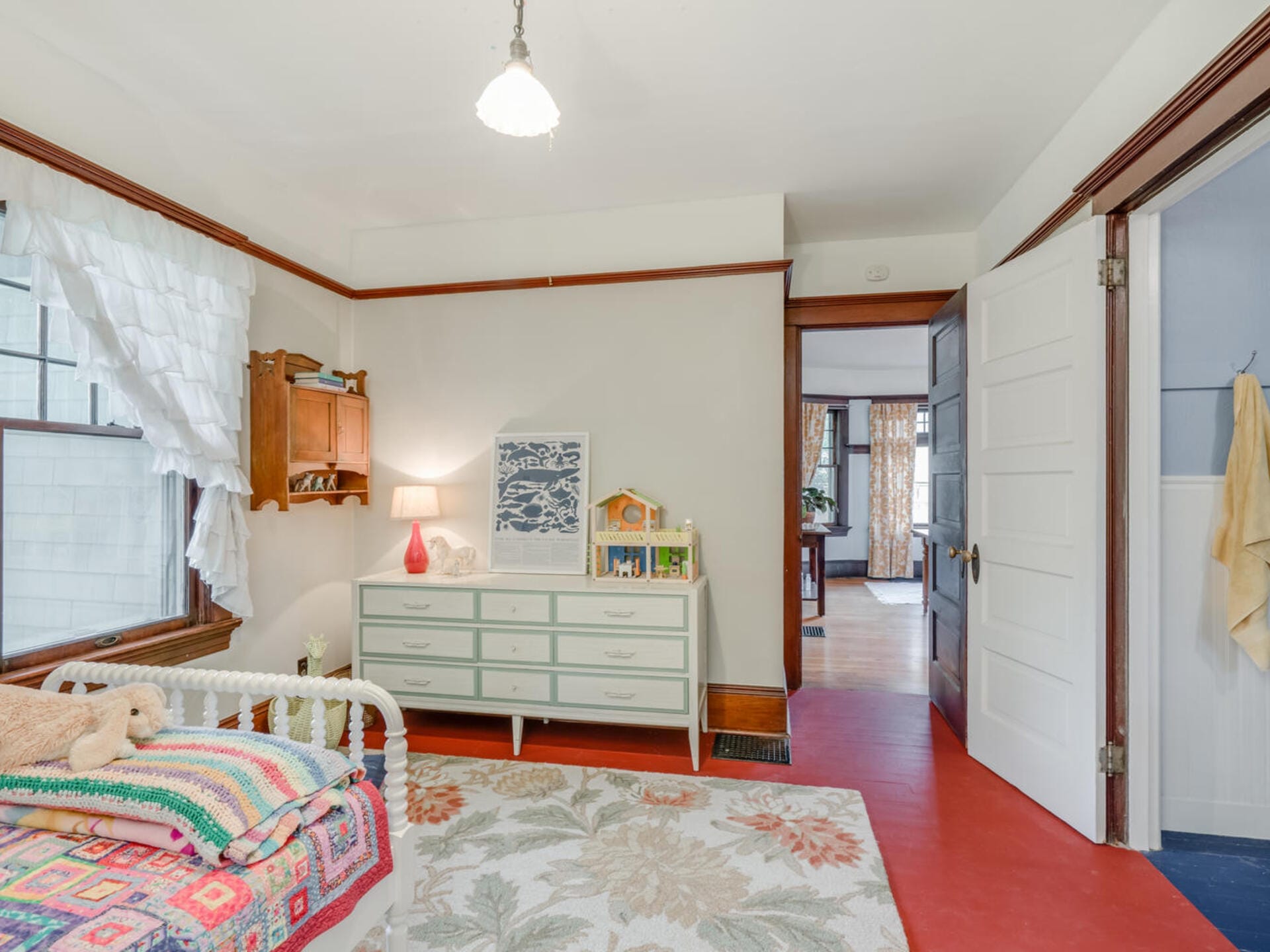 A cozy bedroom with a colorful bed, a patterned rug, and a dresser topped with a lamp and toy house. The walls are light-colored, and a window with sheer curtains brings in natural light. An open door leads to another room.