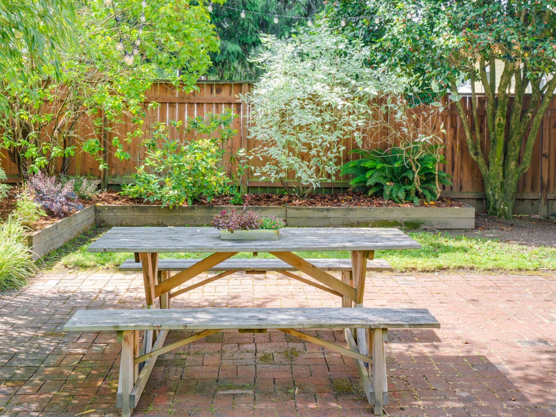 A wooden picnic table and benches are set on a brick patio in a backyard. The area is surrounded by a wooden fence and green foliage, including leafy trees and shrubs, creating a serene and inviting outdoor space.
