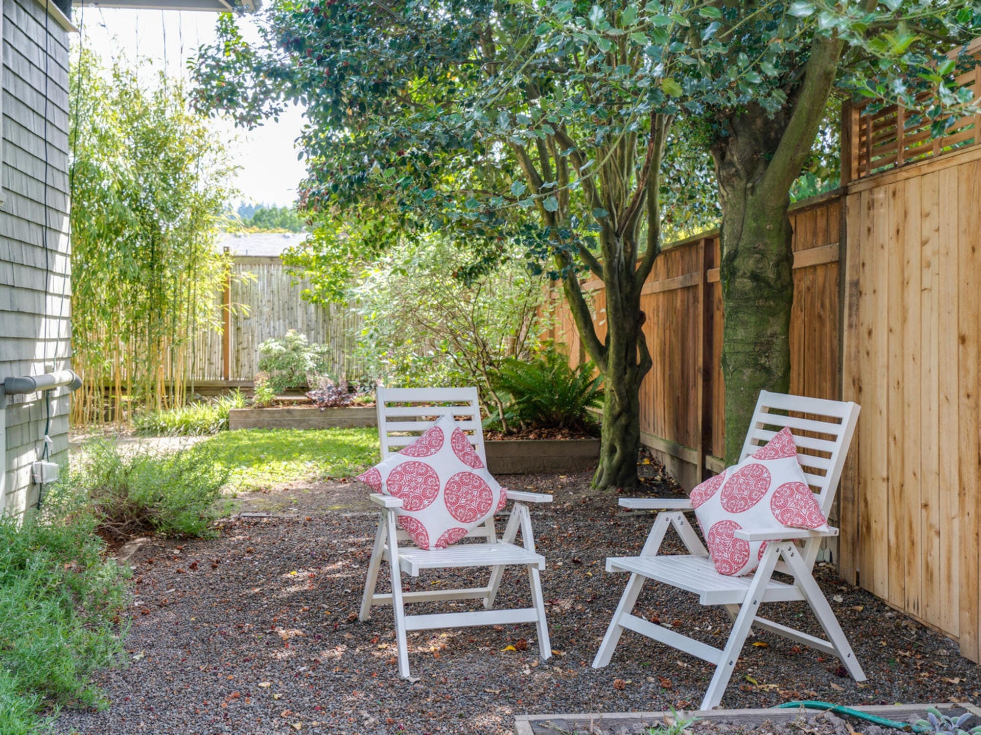 A serene backyard scene with two white chairs placed on gravel. Each chair has a pink-patterned cushion. They sit under the shade of a tree, next to a wooden fence, with greenery and bamboo in the background.