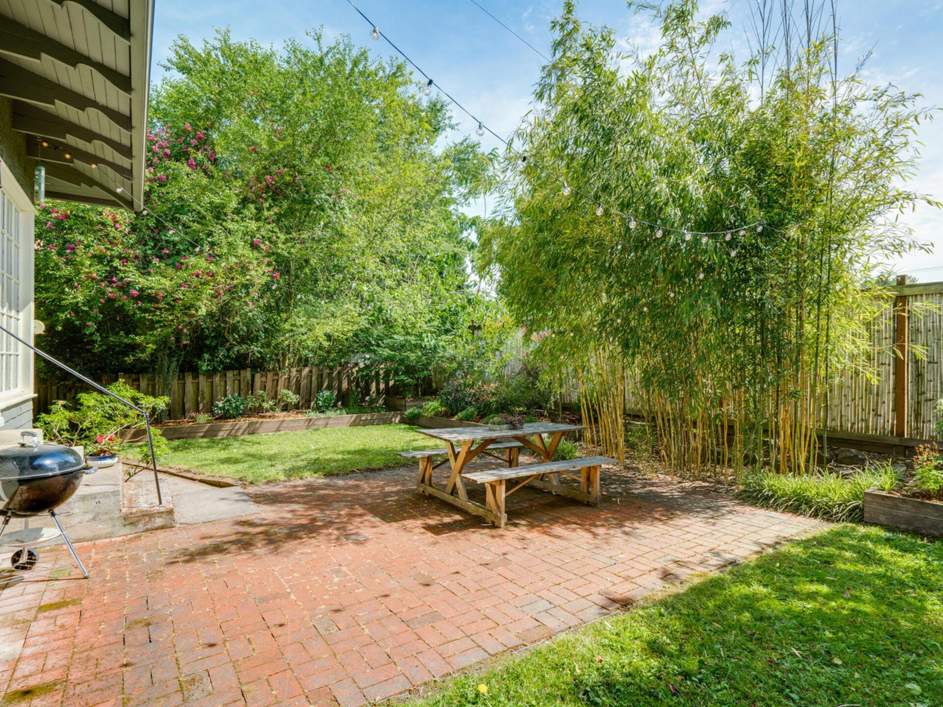 A backyard with a brick patio featuring a picnic table and a charcoal grill. Lush greenery surrounds the area, including bamboo and various trees. String lights are hung above, creating a serene outdoor space.