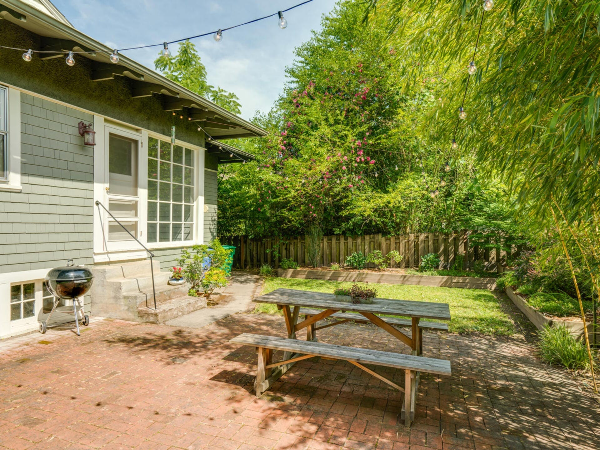 A serene backyard features a wooden picnic table and a charcoal grill on a brick patio. String lights are hung overhead. The space is surrounded by lush greenery and a wooden fence, with a house in the background.