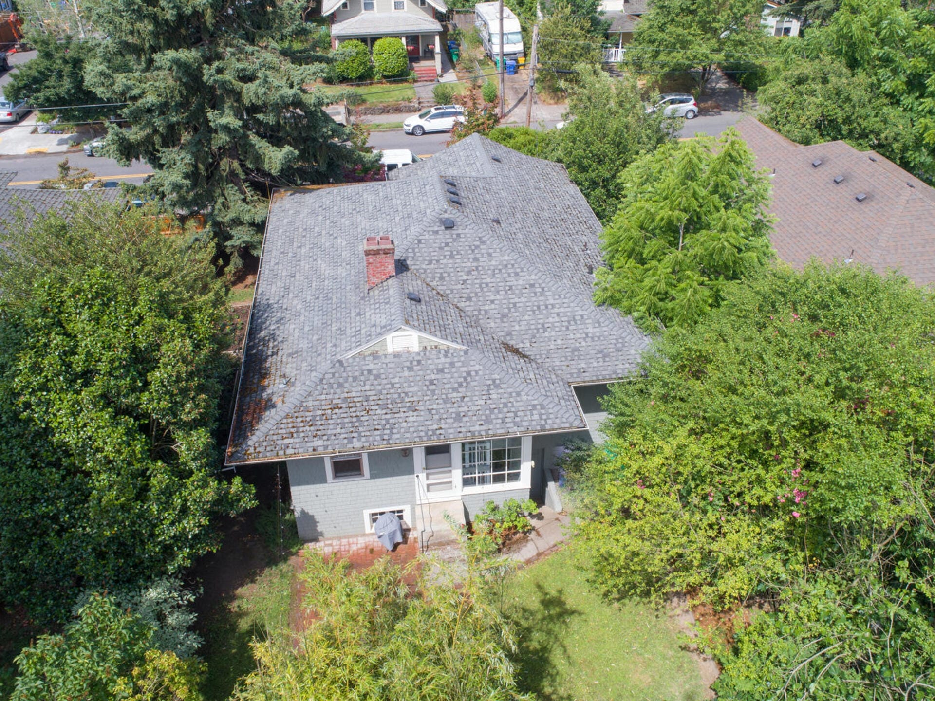 Aerial view of a suburban residential area featuring a house with a gray roof surrounded by lush green trees and a small yard. Other houses and a street with parked cars are visible in the background.