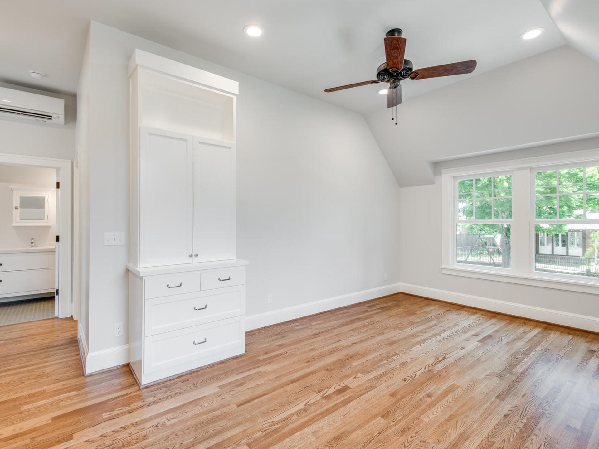 A spacious, empty room in Portland, Oregon features hardwood floors, white walls, and a ceiling fan. A large window floods the space with natural light. Built-in white cabinets and storage drawers line one wall, adding both elegance and functionality.
