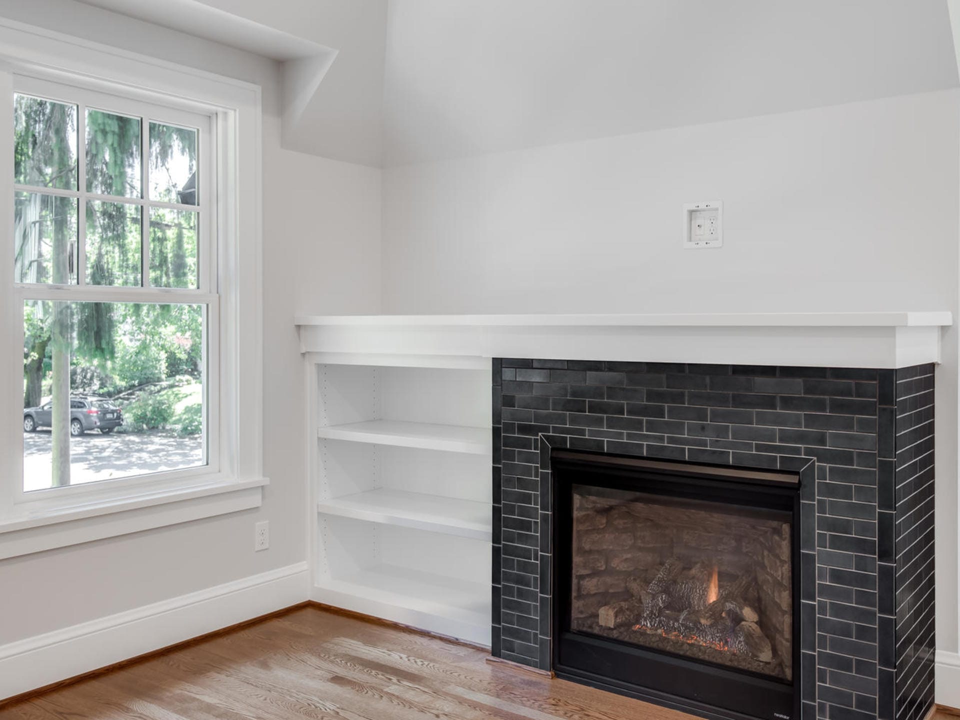 A room in Portland, Oregon features a black-tiled fireplace with a white mantel, adjacent to built-in white shelves. A large window on the left offers a view of greenery outside. The wooden floor complements the neutral wall tones, creating a harmonious and inviting space.