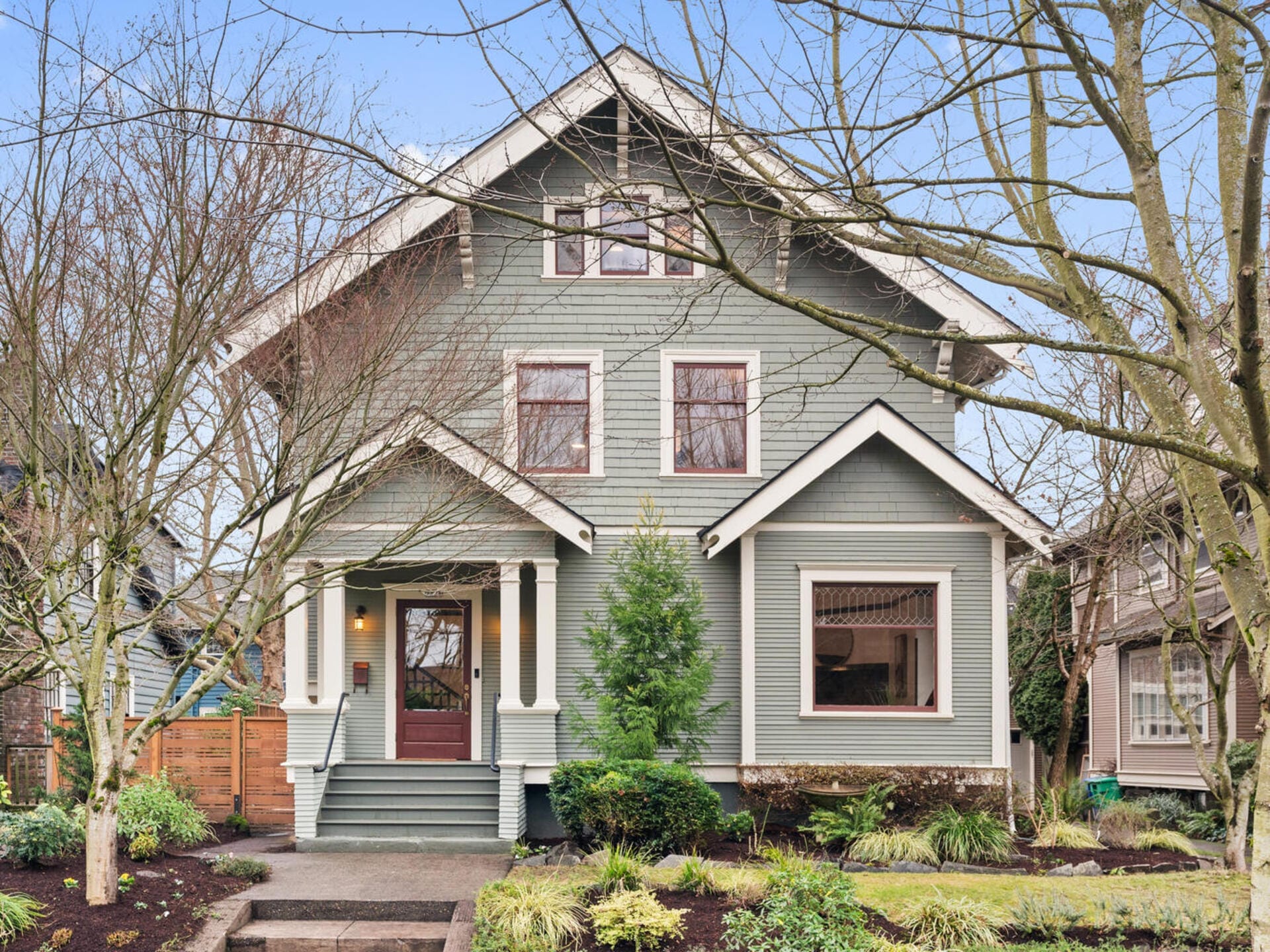 A charming two-story house with a gray exterior and white trim, surrounded by bare trees and lush landscaping. The home features a covered front porch and multiple windows, creating a welcoming appearance. A clear sky is visible in the background.