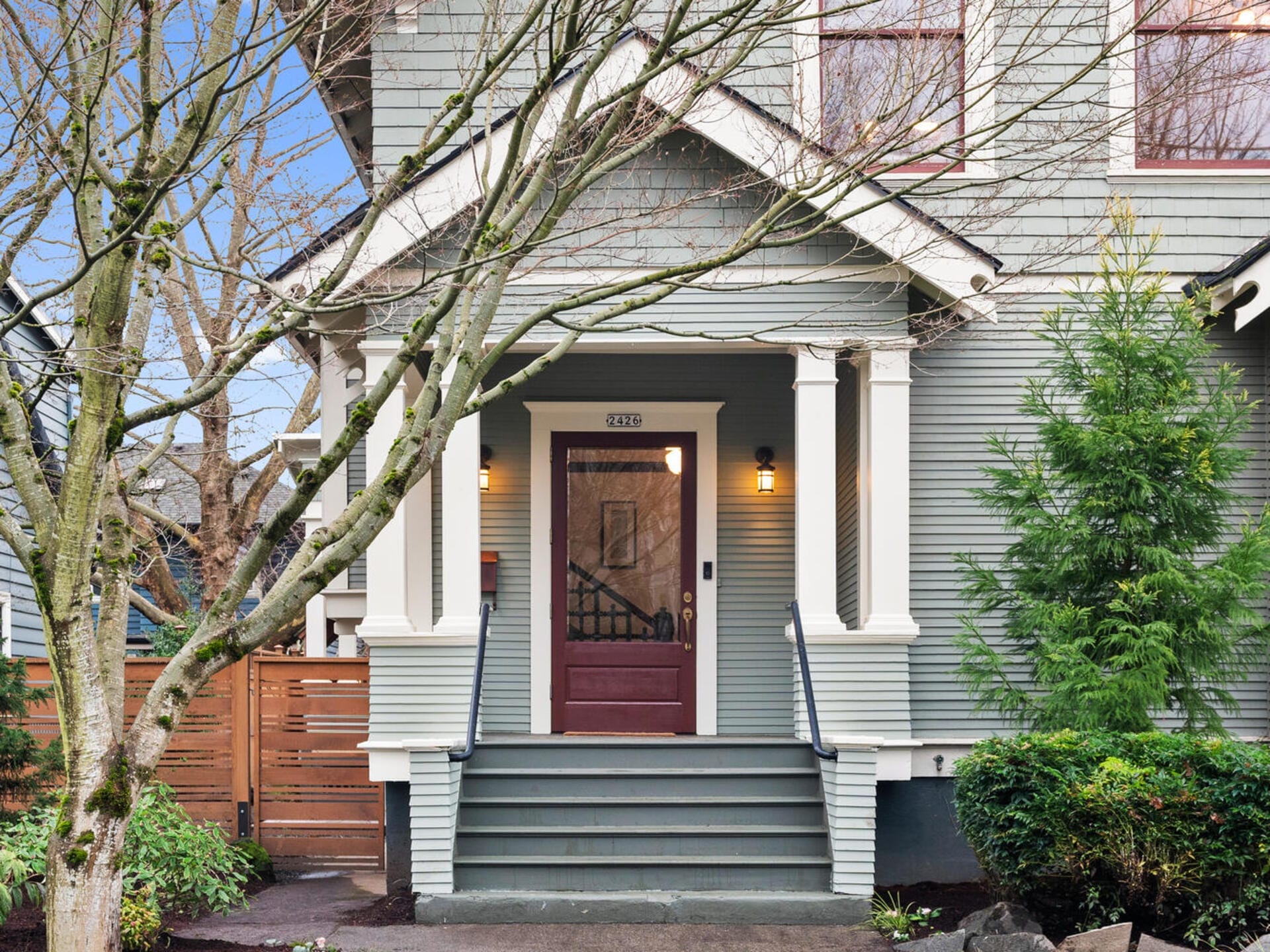 Front view of a two-story house with pale green siding and white trim. There are steps leading to a maroon front door, framed by columns. Trees and shrubs decorate the front yard, and a wooden fence is visible on the left.