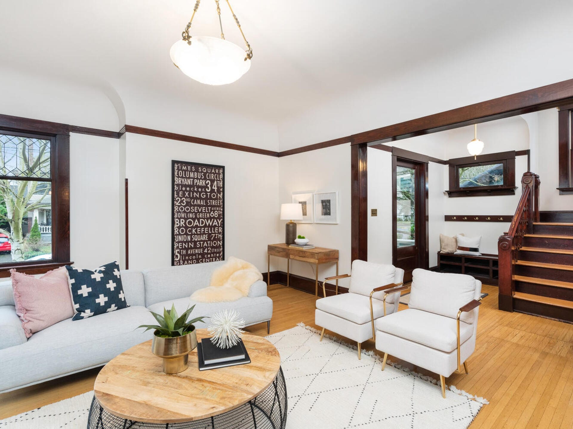 A cozy living room with hardwood floors, featuring a light gray sofa, two white chairs, and a round wooden coffee table. Art on the wall and a staircase in the background add to the elegant, inviting atmosphere.