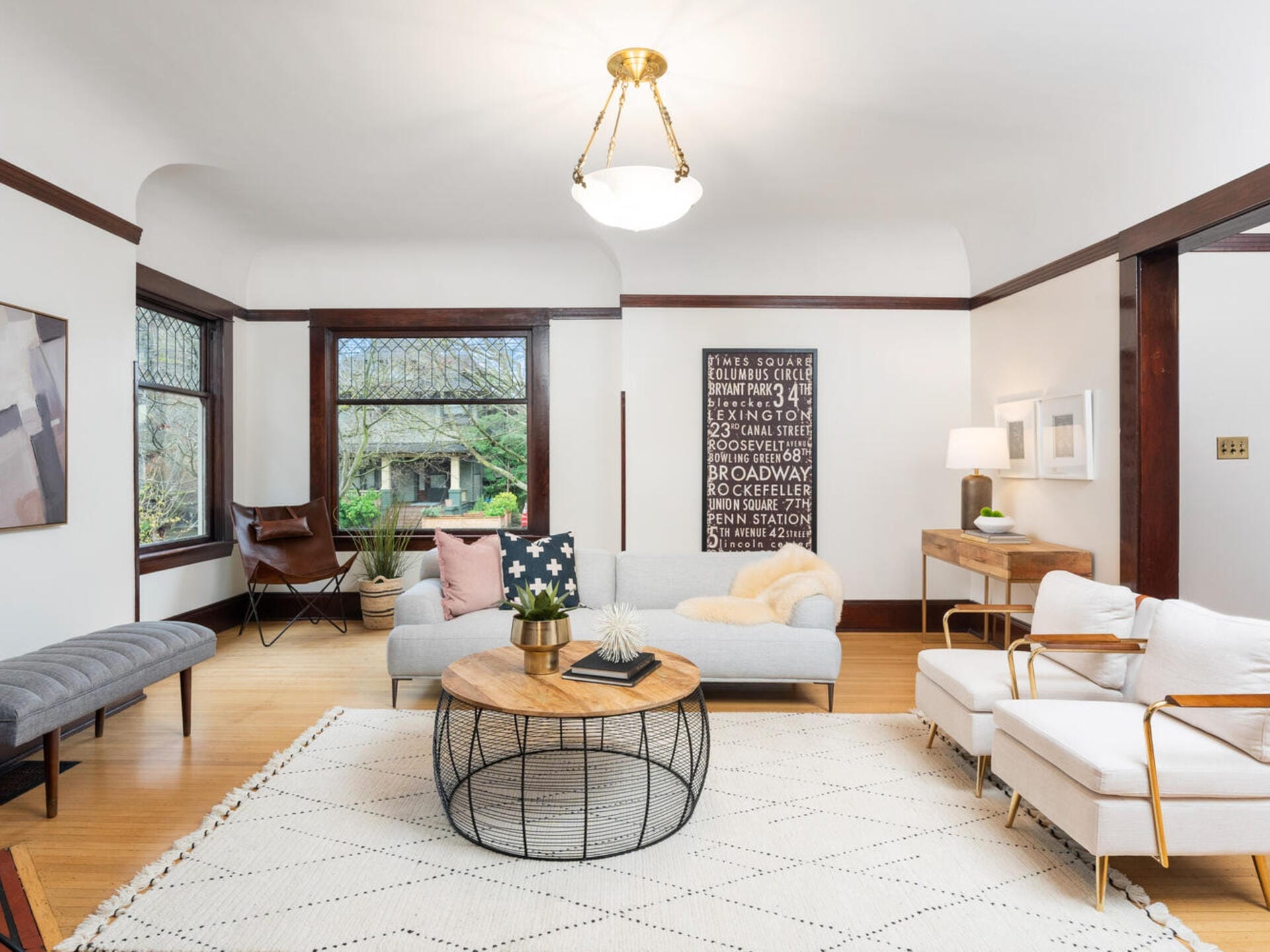 A bright, spacious living room with light-colored walls and hardwood floors. It features a white sofa, a round coffee table, a floor lamp, a large window with natural light, and a modern wall art piece. A cozy chair and bench complete the setting.
