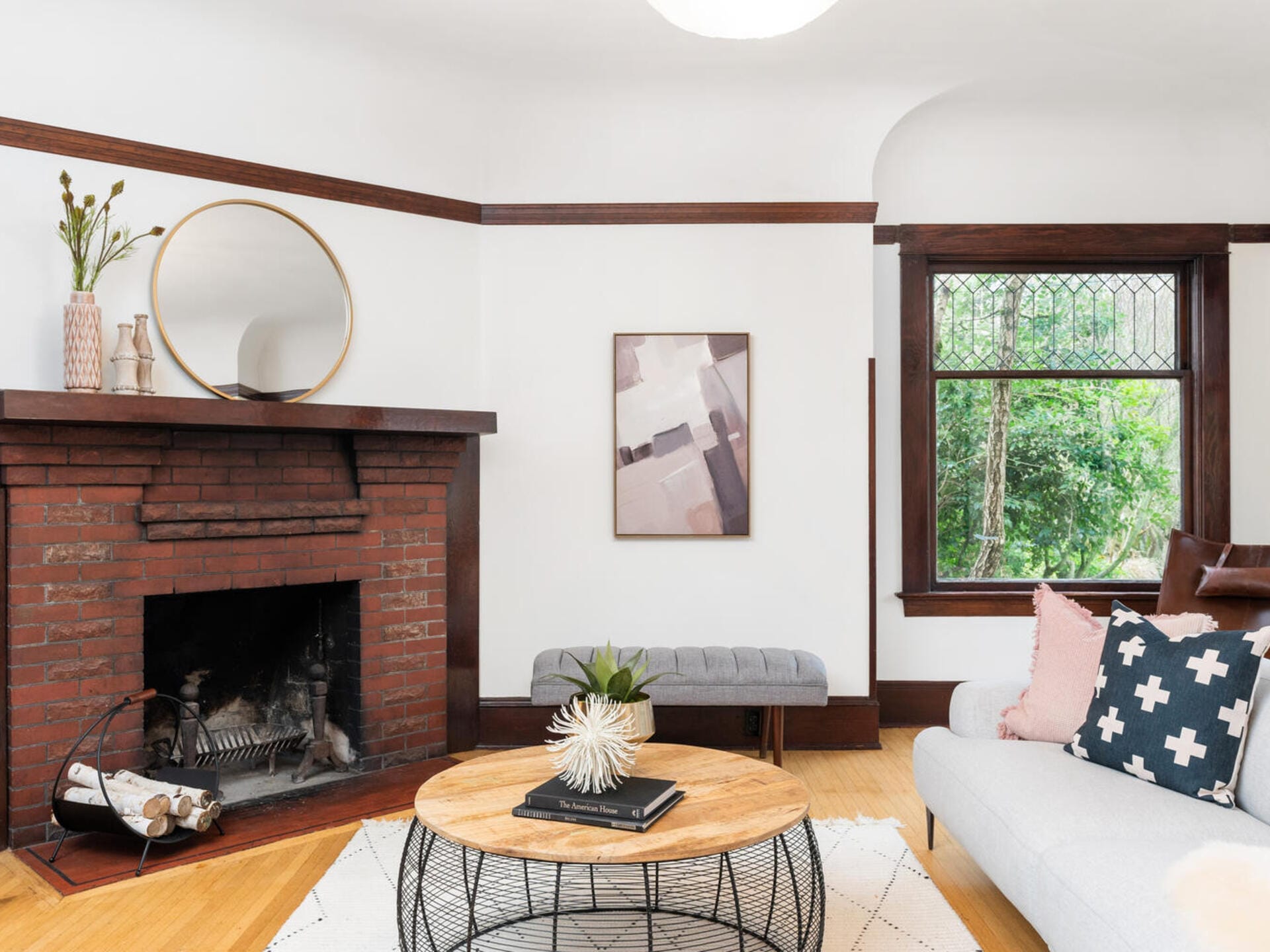 A cozy living room with a brick fireplace, round mirror, and decorative vases on the mantel. A light gray sofa with patterned pillows sits next to a large window. A round wooden coffee table rests on a geometric rug.
