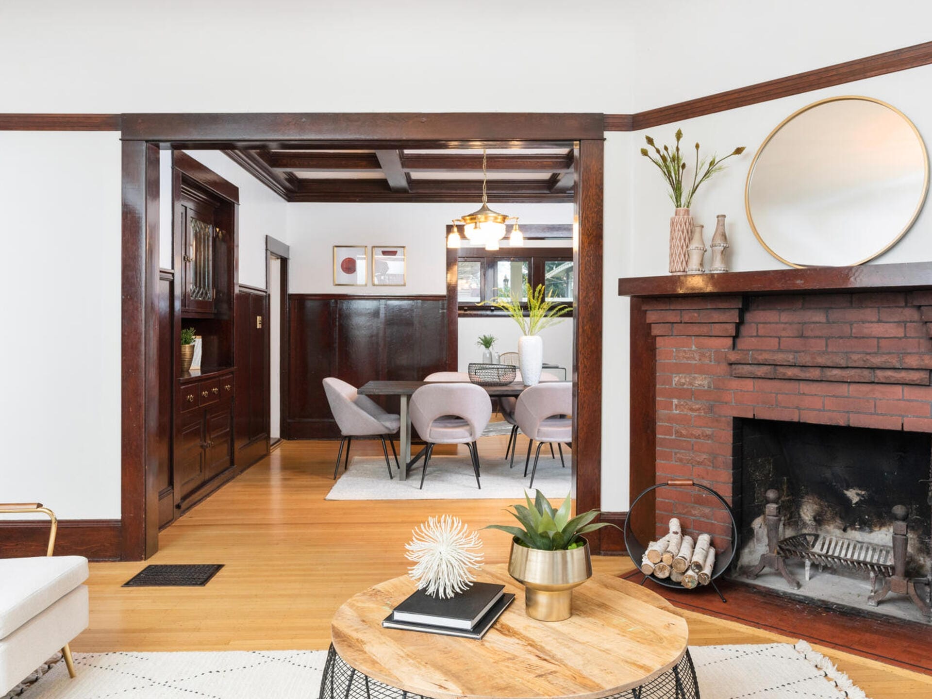 A cozy living room with a brick fireplace on the right, a round coffee table with decorations in the center, and an archway leading to a dining area with a wooden floor, purple chairs, and a round chandelier.