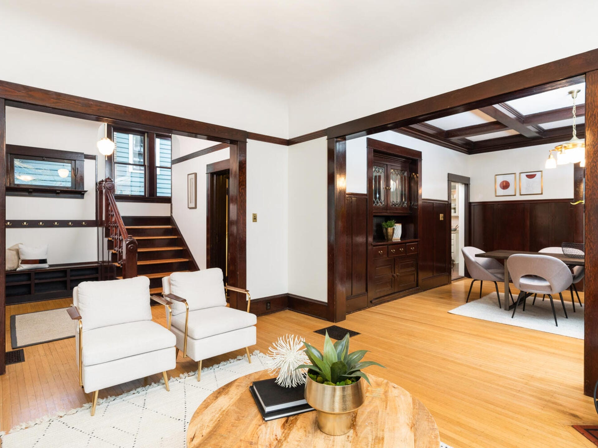 A stylish open living space with two white armchairs and a wooden coffee table in the foreground. The room features dark wood trim, a dining area with a round table and chairs, and a staircase leading upstairs. Artwork hangs on the walls.