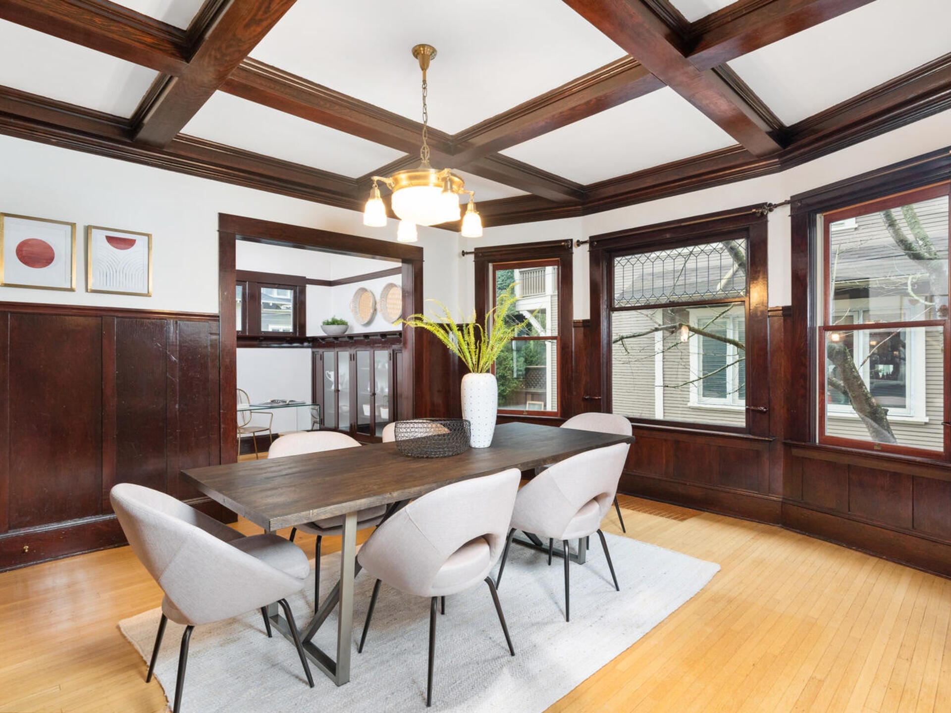 A dining room with a dark wooden table and six modern chairs. The room features a wooden coffered ceiling and wainscoted walls. Large windows allow natural light in, and three framed artworks are on the wall. A plant centerpiece is on the table.