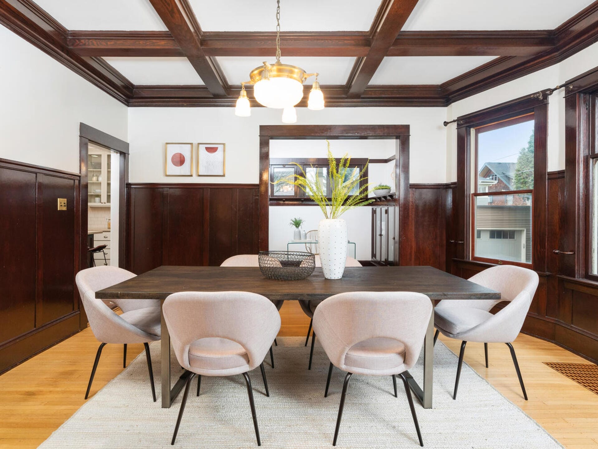 A dining room with a dark wooden coffered ceiling, light beige chairs, and a long rectangular table. The walls have dark wood paneling, and three artworks hang above a sideboard. A vase with yellow flowers decorates the table. Large windows bring in light.