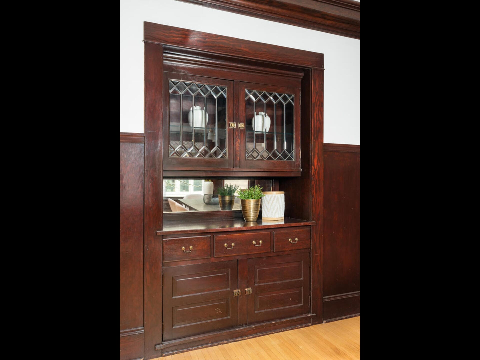 A built-in wooden cabinet with glass doors and a mirror is set against a white wall. The cabinet holds potted plants and decorative items. It features drawers and lower storage compartments, with intricate wood detailing and brass handles.