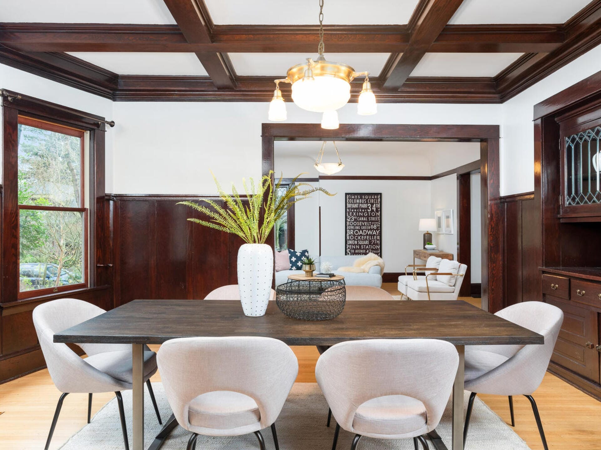 A modern dining room with a wooden table, six light gray chairs, and a large white vase with green foliage as the centerpiece. Dark wood paneling accents the room, with a view into a cozy living area. A chandelier hangs overhead.