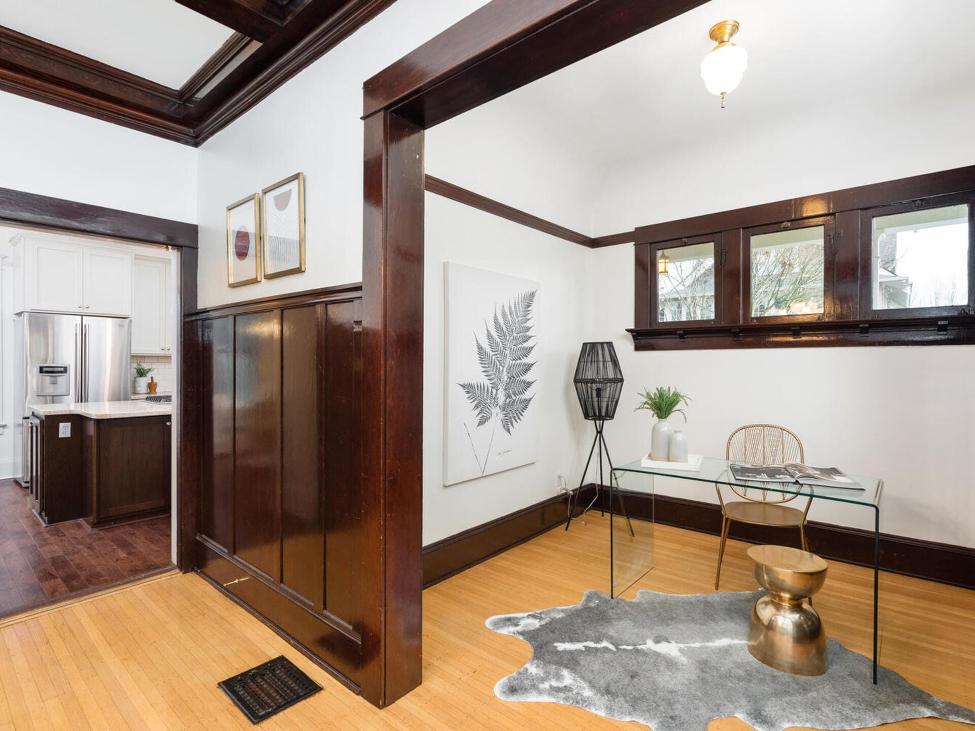 A stylish room features a glass desk with a decorative plant and lamp, a fur rug, and a golden stool. Wood-paneled walls and open passage lead to a kitchen with stainless steel appliances and white cabinets. A fern artwork adorns the wall.
