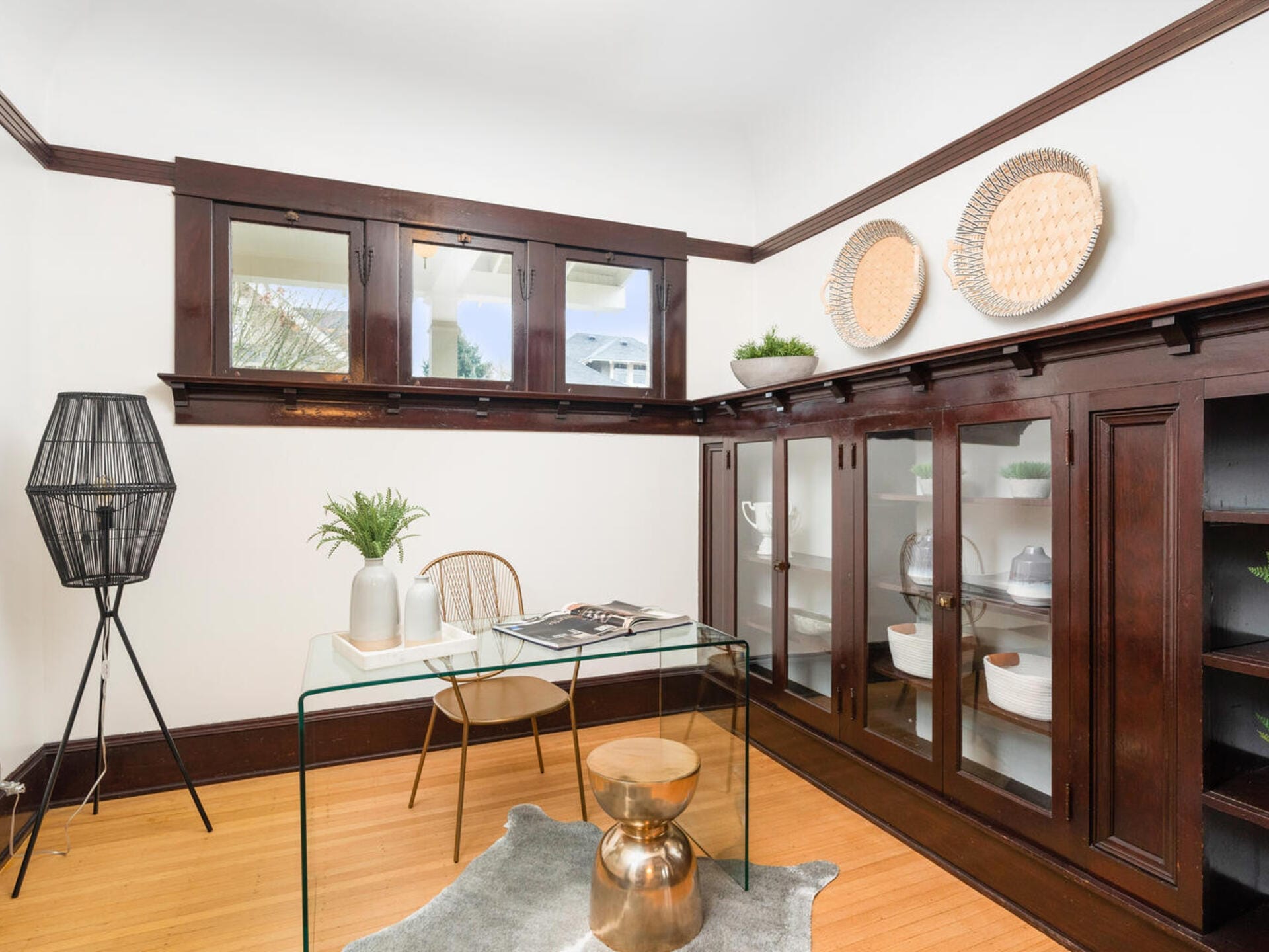 A cozy home office with dark wood built-in cabinets and white walls. The room features a glass desk with a plant and book, a modern black floor lamp, a gold stool, and two decorative plates on the wall. A cowhide rug adds texture on the floor.