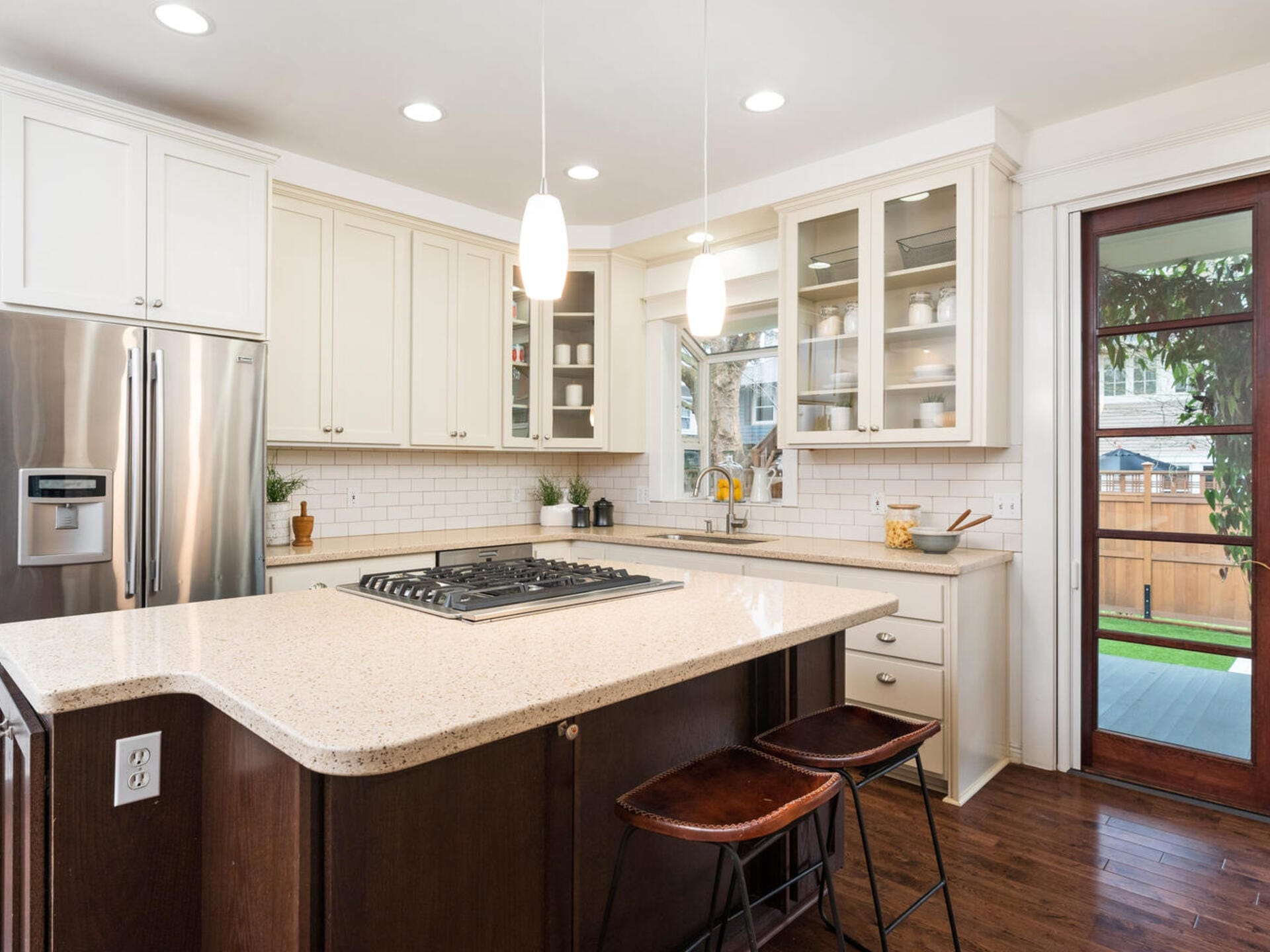 Modern kitchen with white cabinets, stainless steel appliances, and a large island featuring a built-in stove. Two pendant lights hang above the island, with two wooden stools. A glass door leads to an outdoor area with a view of greenery and a fence.