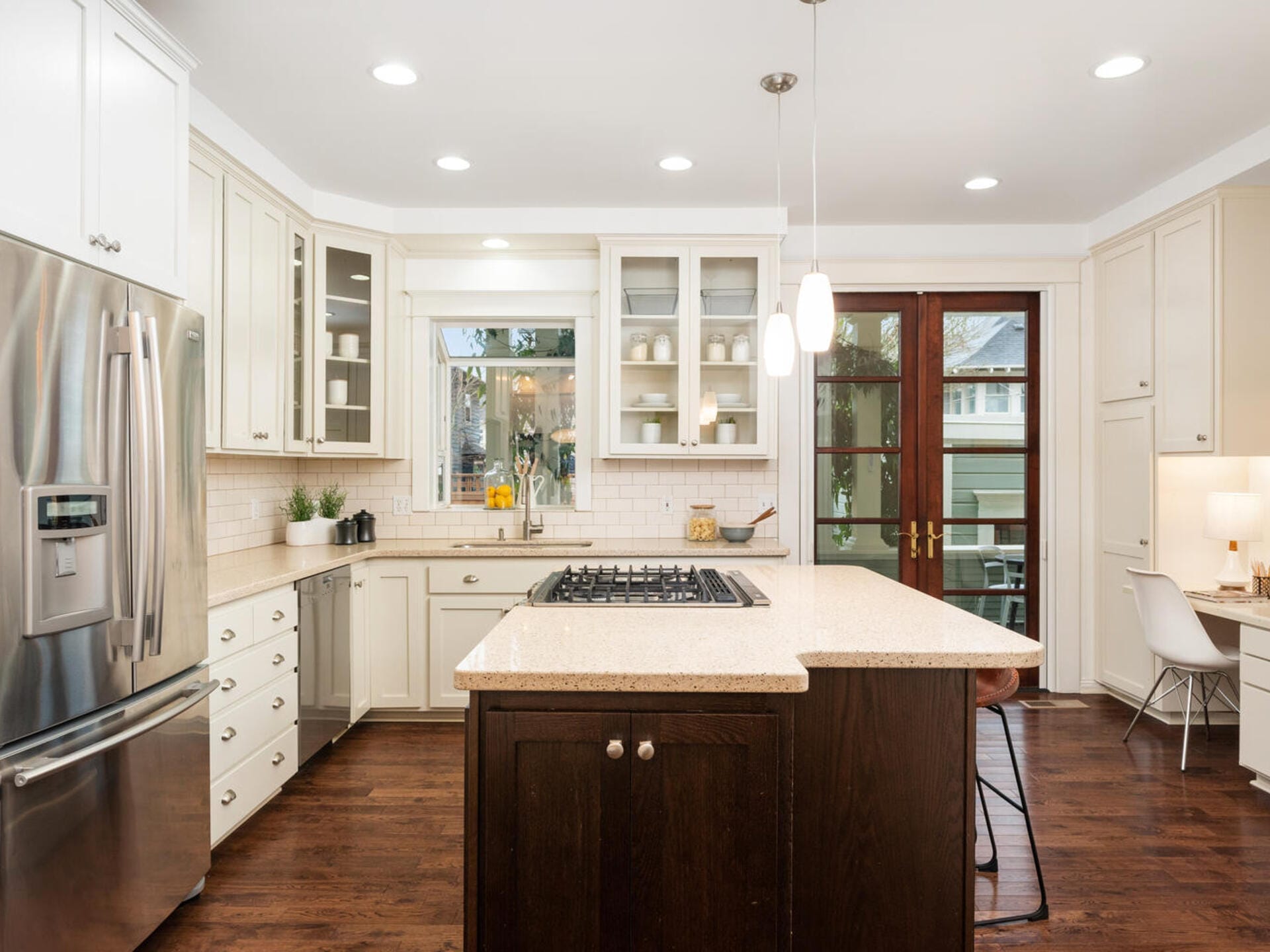A modern kitchen with white cabinets, a large island with a stovetop, and stainless steel appliances. Dark wooden flooring contrasts with light walls. A small desk area is in the corner near double glass doors leading outside. Pendant lights hang above.