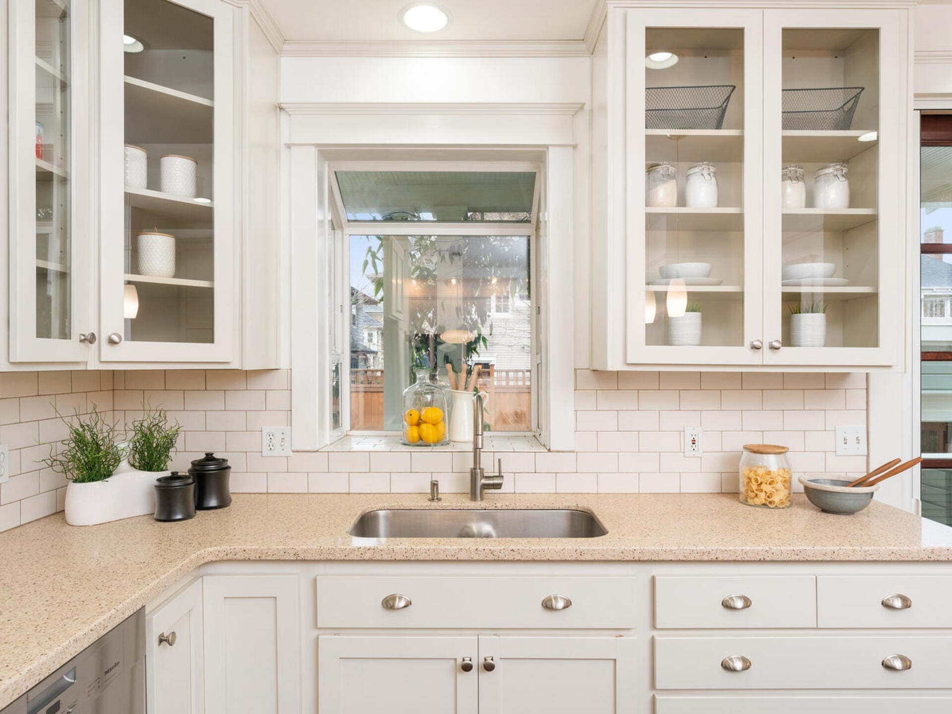 A bright kitchen with white cabinets, glass-front doors, and a beige countertop. A sink is centered under a window with potted herbs and jars on either side. Lemons are in a bowl near the window, and light enters from the outside.
