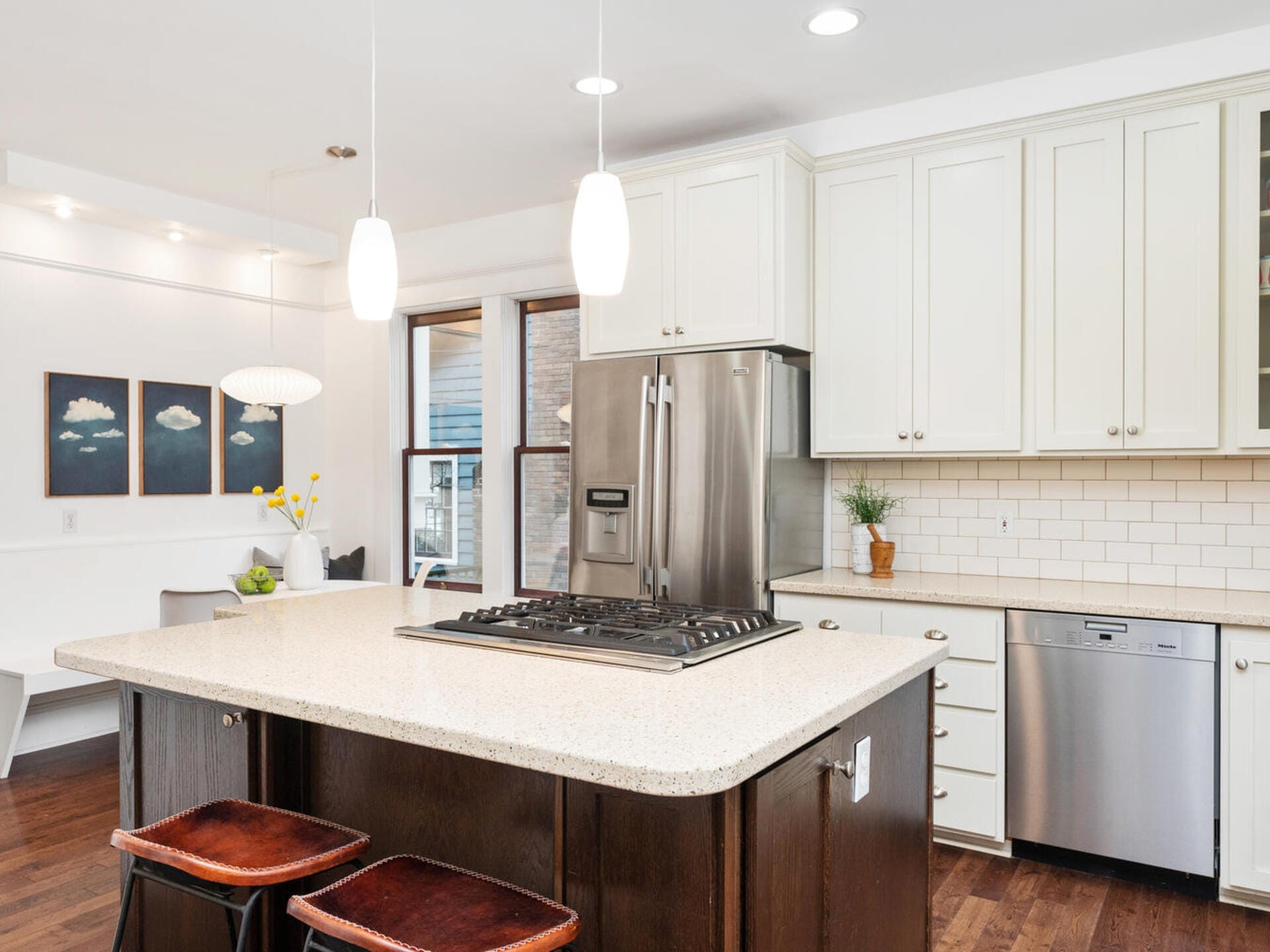 A modern kitchen with white cabinets, stainless steel appliances, and a gas stove on a large island. Two pendant lights hang above, with wood floors and seating at the island. A small plant and kitchen decor add a cozy touch.