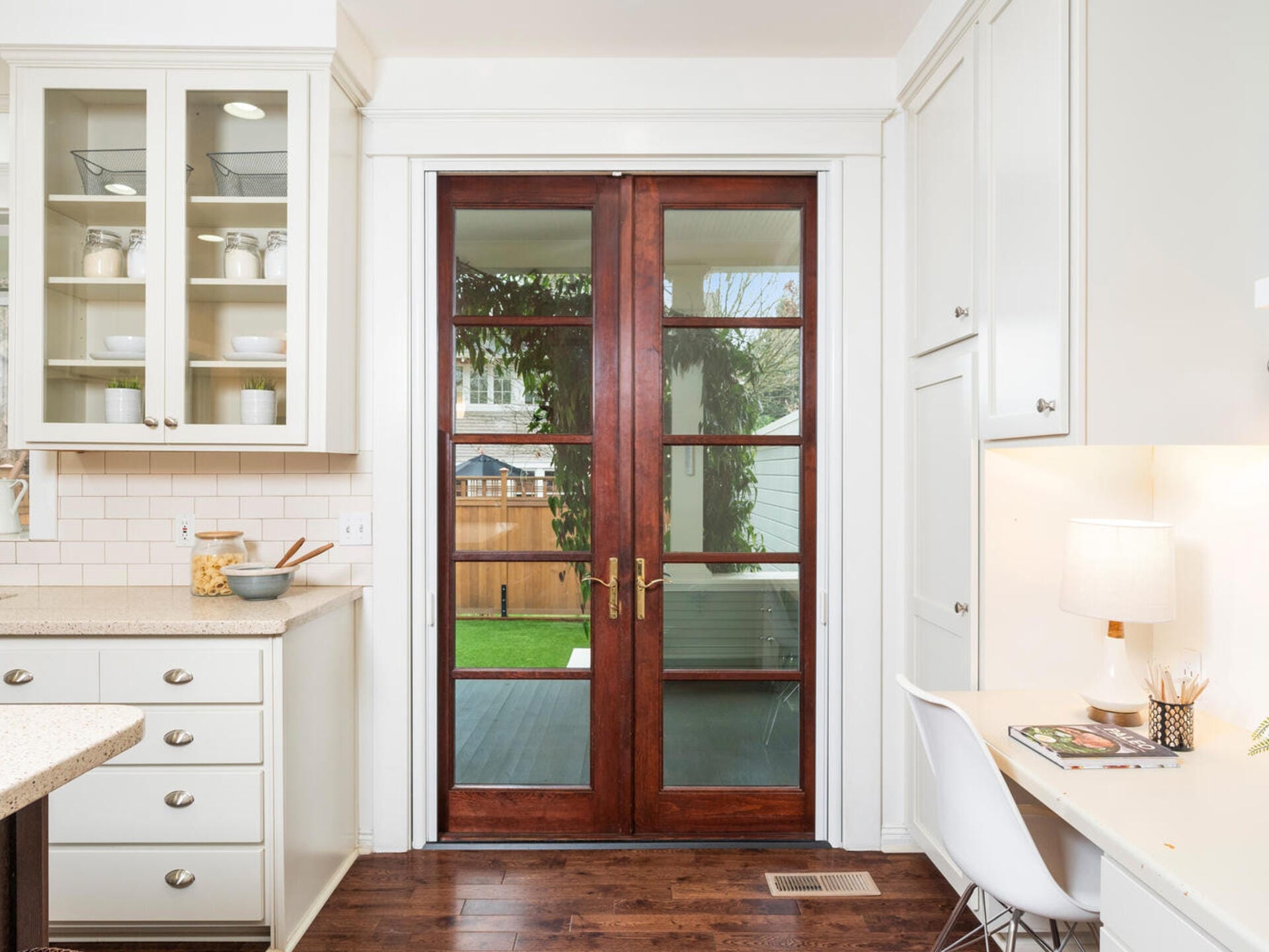 A kitchen with white cabinets and a counter is adjacent to a small office space with a white desk and chair. Wooden double doors with glass panes lead outside to a backyard with a fence and greenery.