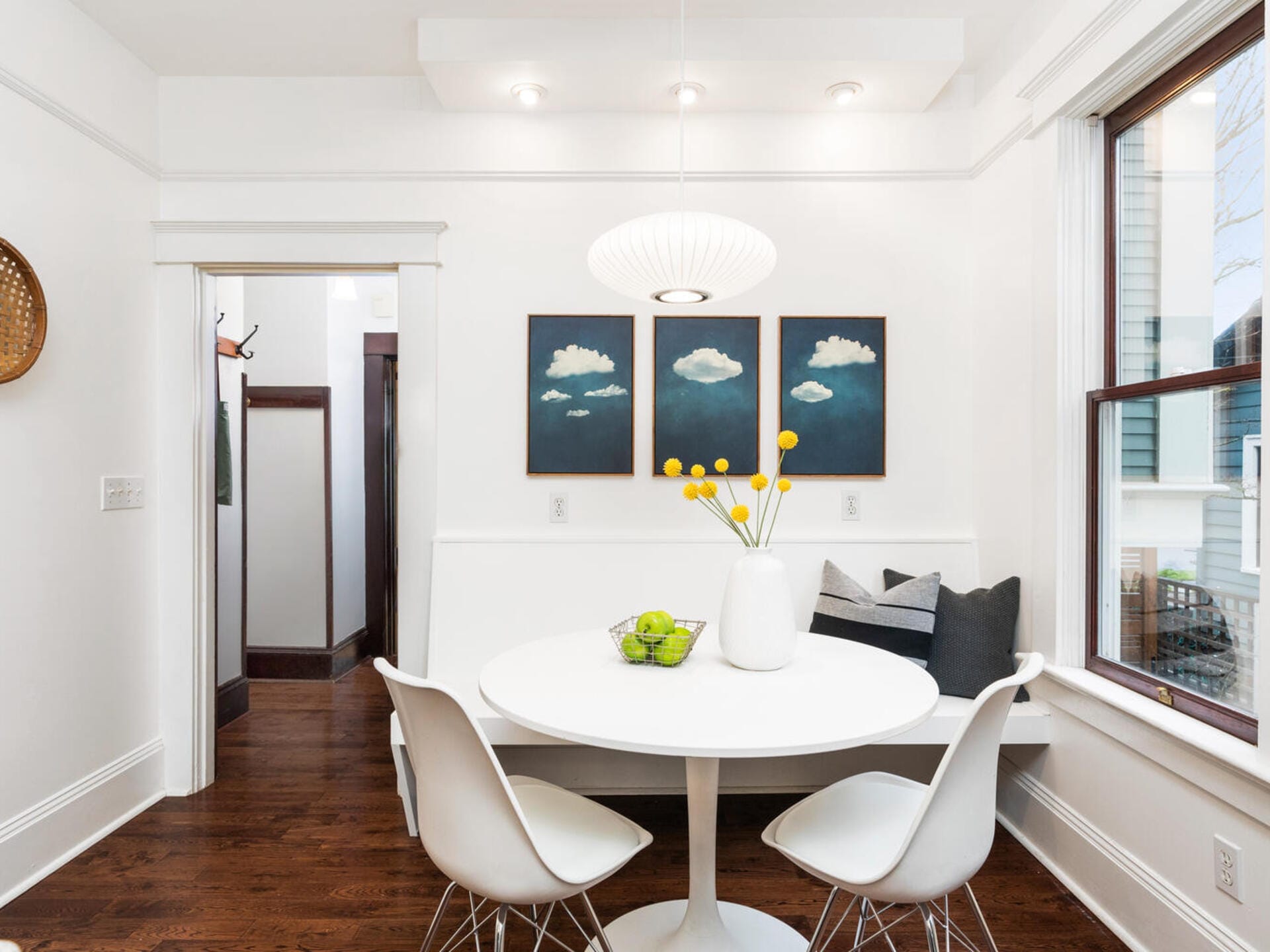 A modern dining room with a round white table and three white chairs. The table has a bowl of green apples and a vase with yellow flowers. Three cloud paintings hang on the wall. A pendant lamp is above the table, and a window is on the right.