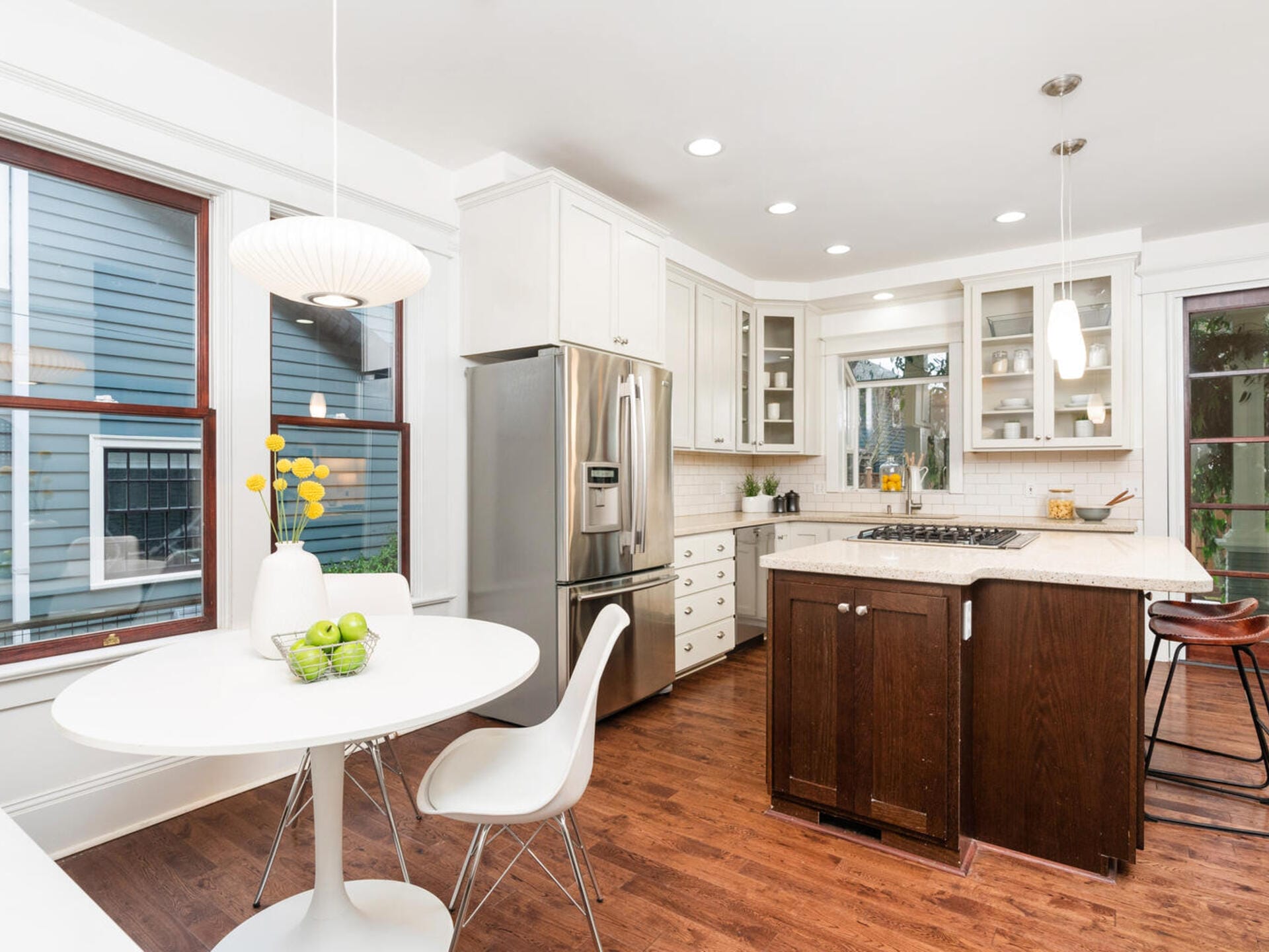 A modern kitchen with white cabinetry, a stainless steel refrigerator, and a central island with a dark wood base. There’s a round white dining table with a vase of yellow flowers and green apples. Wooden floor and pendant lights complete the space.