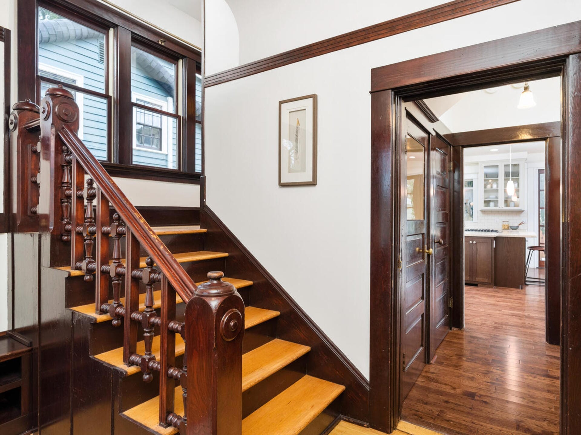 A hallway with wooden flooring leads to a well-lit kitchen. A wooden staircase with carved railings is on the left. White walls contrast with dark wood trims and doors. A framed picture hangs on the wall above the stairs.