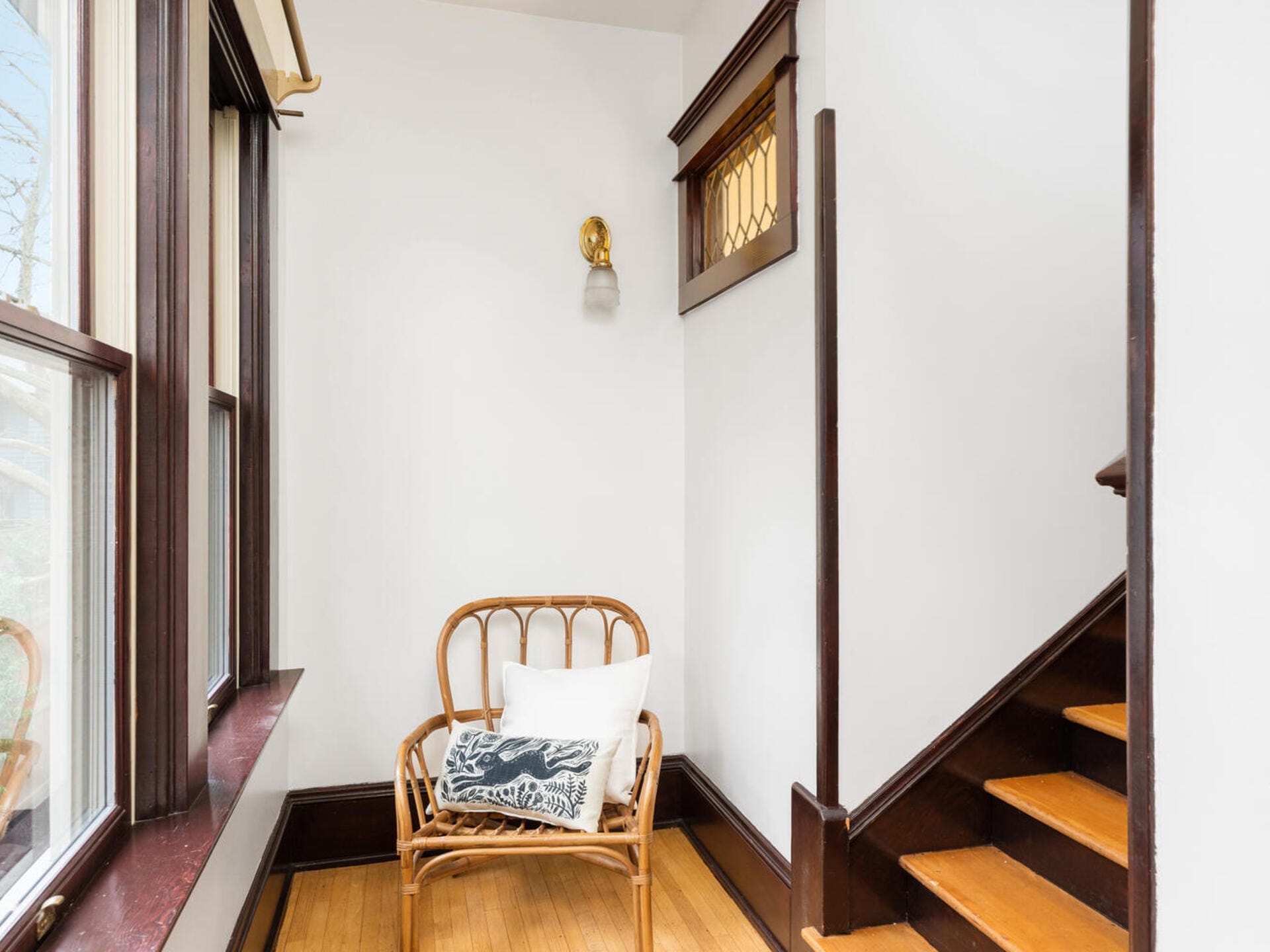 A cozy nook with a wooden chair and cushion, next to a staircase with wooden steps and railing. A small wall light is above the chair, and natural light streams through the windows, illuminating the white walls and warm wood tones.