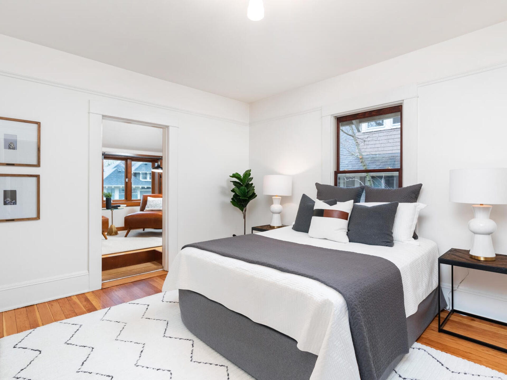 A bright bedroom with a neatly made bed featuring gray and white bedding. A large window lets in natural light. Two bedside tables hold lamps. Framed artwork hangs on one wall, and a green plant adds a touch of color. A second room is visible.