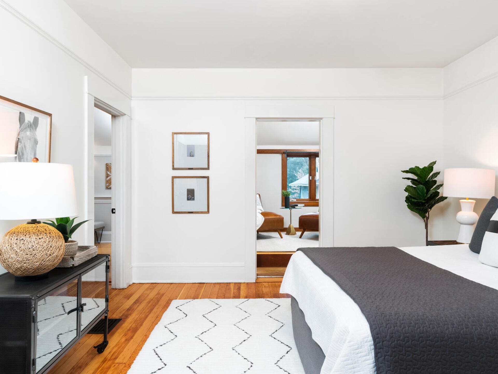A modern bedroom with white walls, a bed with gray and white bedding, and wooden floors. A large plant and two framed artworks decorate the space. A glimpse of a sitting area with a window and chairs is visible through an open door.