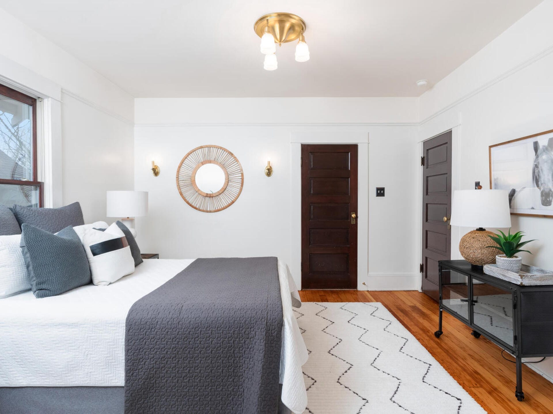 A bright bedroom features a neatly made bed with gray and white bedding, a round mirror with a decorative frame on the white wall, dark wooden doors, a light fixture, a gray rug with a zigzag pattern, and a black console table with decor items.