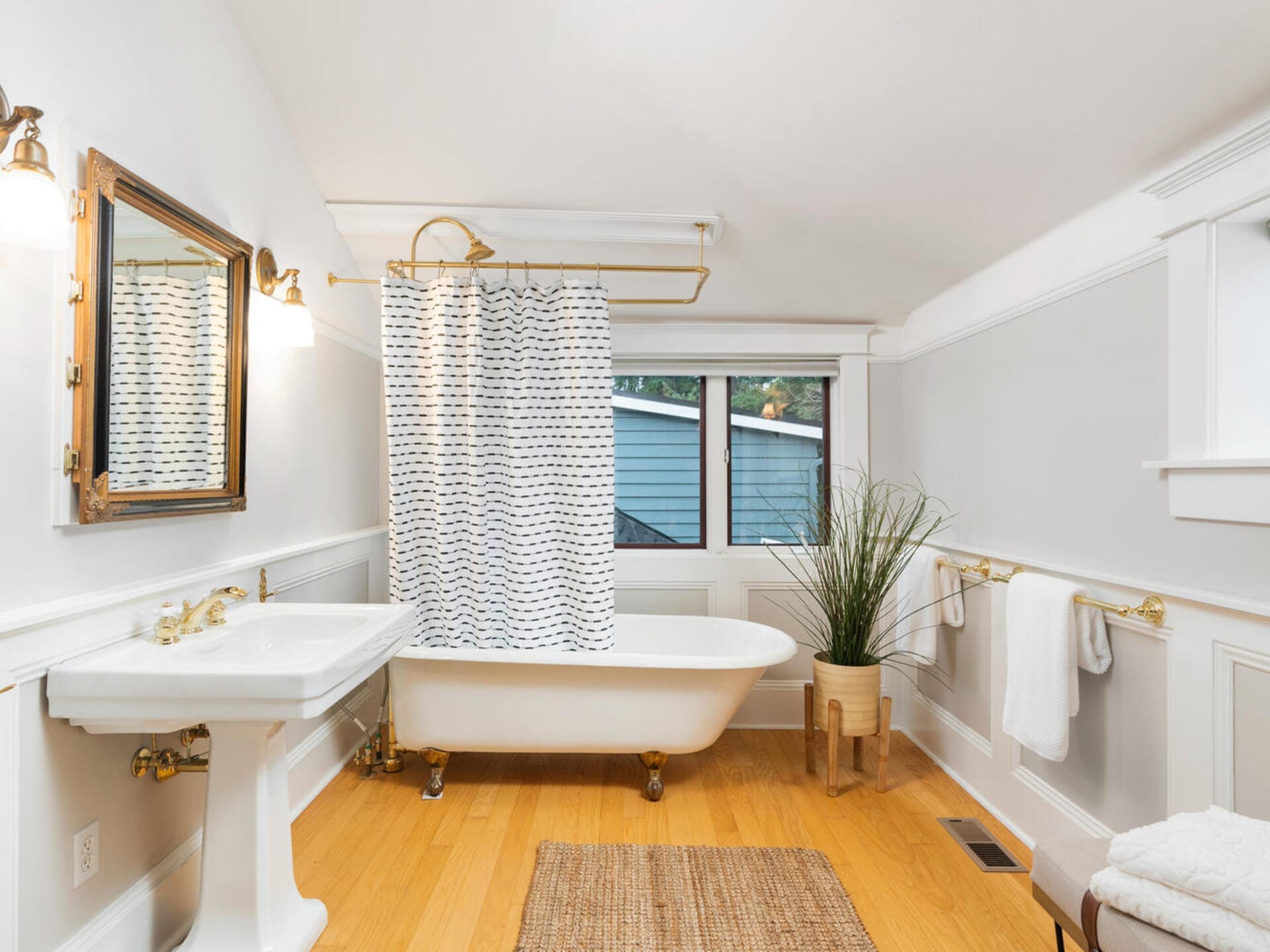 A bright bathroom with a white clawfoot tub and a black-and-white patterned shower curtain. Theres a white sink with a mirror above it, a potted plant, and towels on racks. The floor is wooden, and a window lets in natural light.
