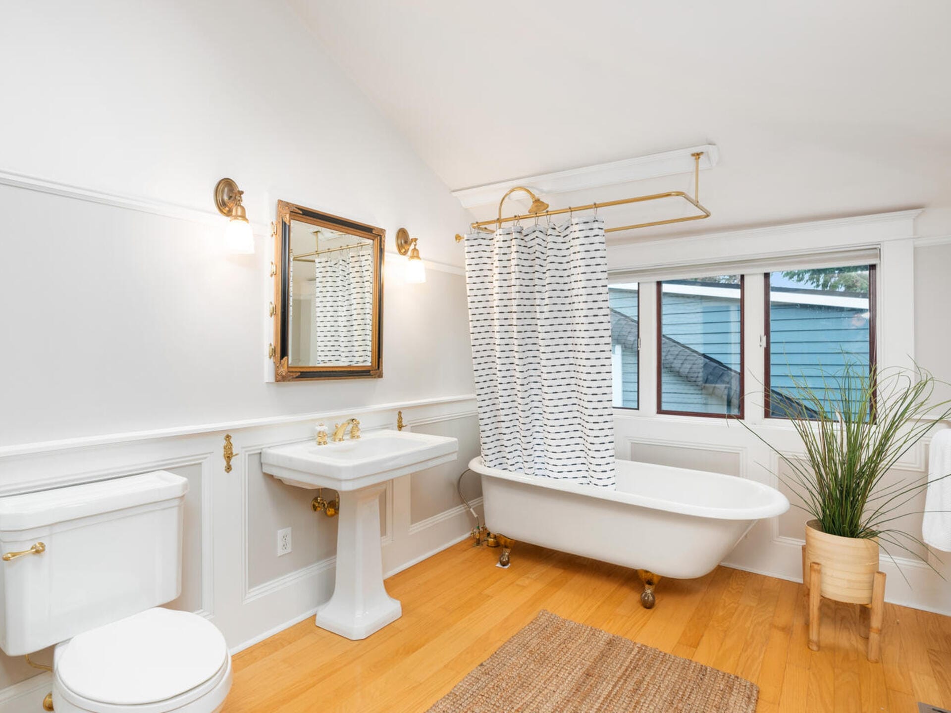 A bright bathroom with a freestanding clawfoot bathtub, a shower curtain, and a window. It features a white sink with a mirror, a toilet, a potted plant, wood floors, and wall-mounted lights. A woven rug is on the floor.