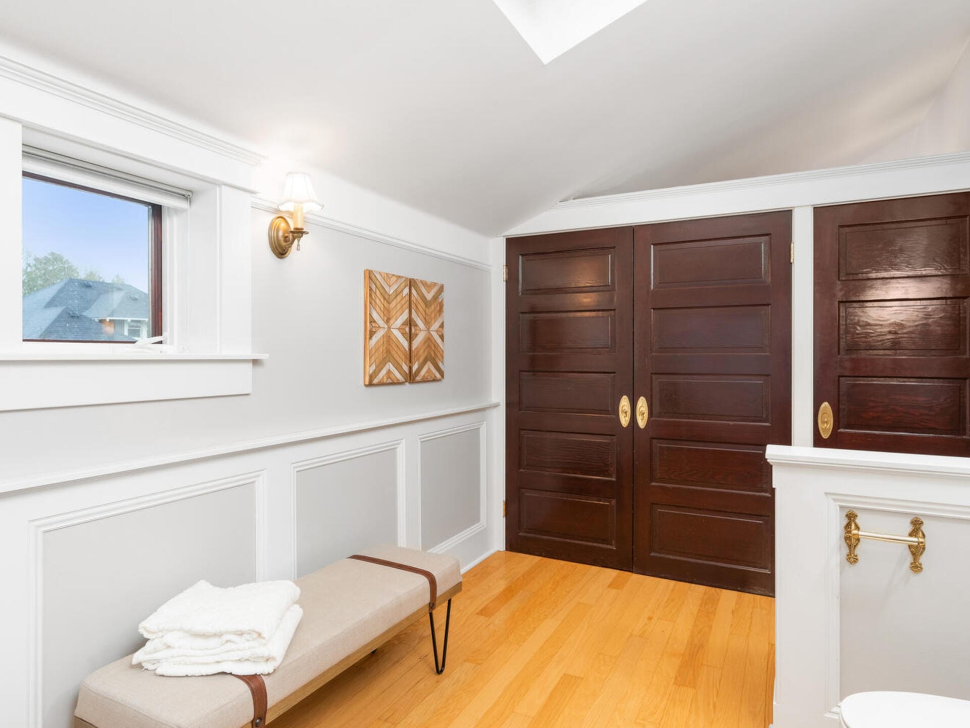 A bright, minimalist room features a skylight, a white paneled wall, a window, and two dark wooden doors. There is a beige bench with folded towels, a geometric wooden wall art piece, and a brass wall sconce. Light wood flooring adds warmth.